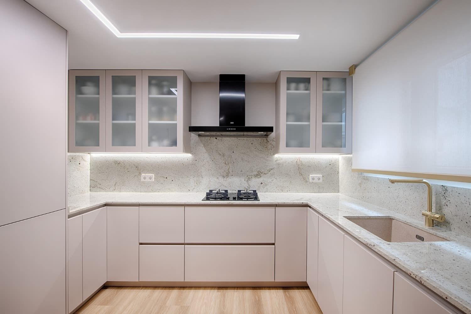 Cocina luminosa y minimalista en forma de L con armarios de tonos claros, encimeras de mármol y campana extractora negra.