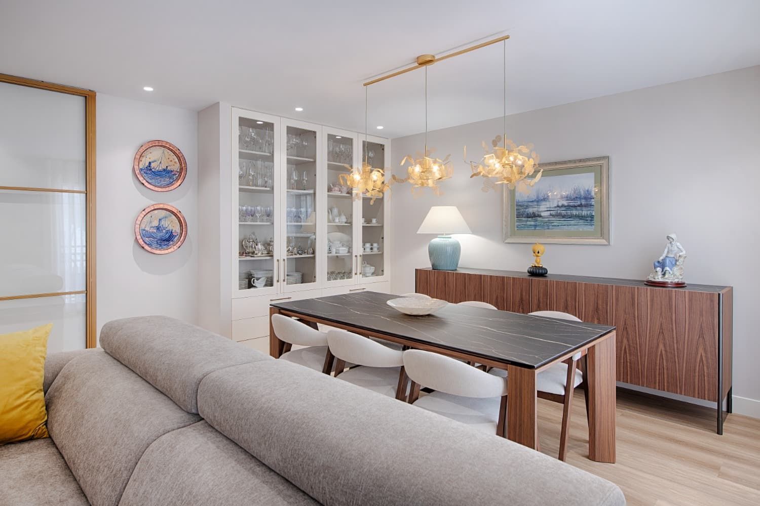 Comedor y sala de estar luminosos con mesa de madera, sofá gris, vitrina y cuadros en la pared.