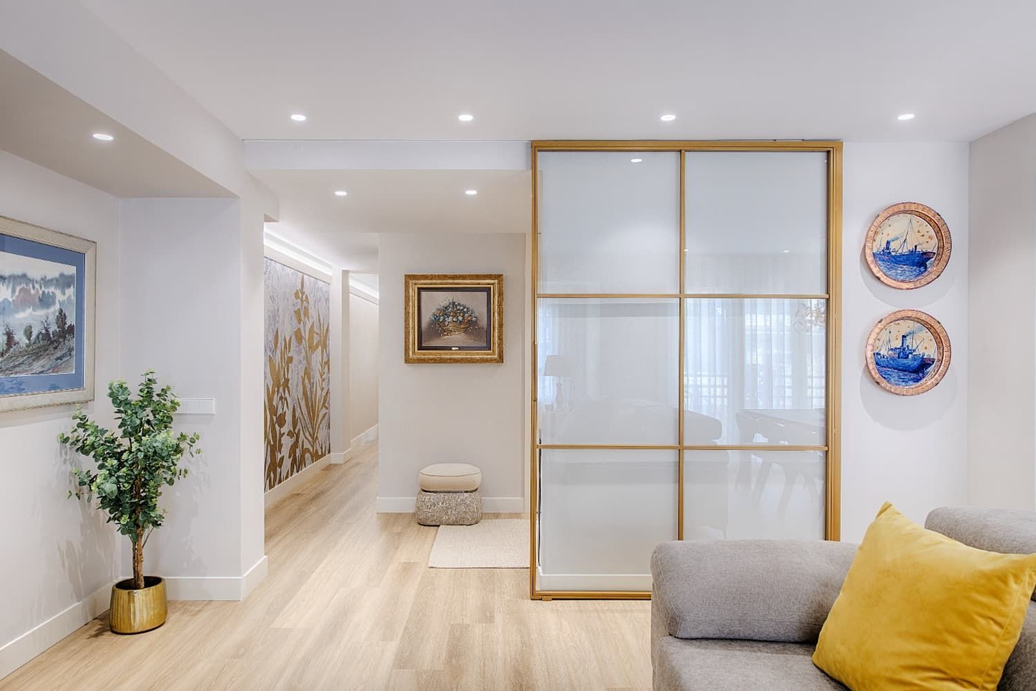 Luminoso salón moderno con suelos de madera clara, separador corredero con marco dorado y sofá gris con cojín amarillo.