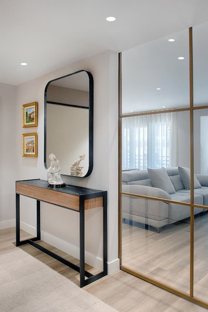 Pasillo moderno con mesa consola, espejo arqueado y pared de cristal con marco dorado que da a una sala de estar.