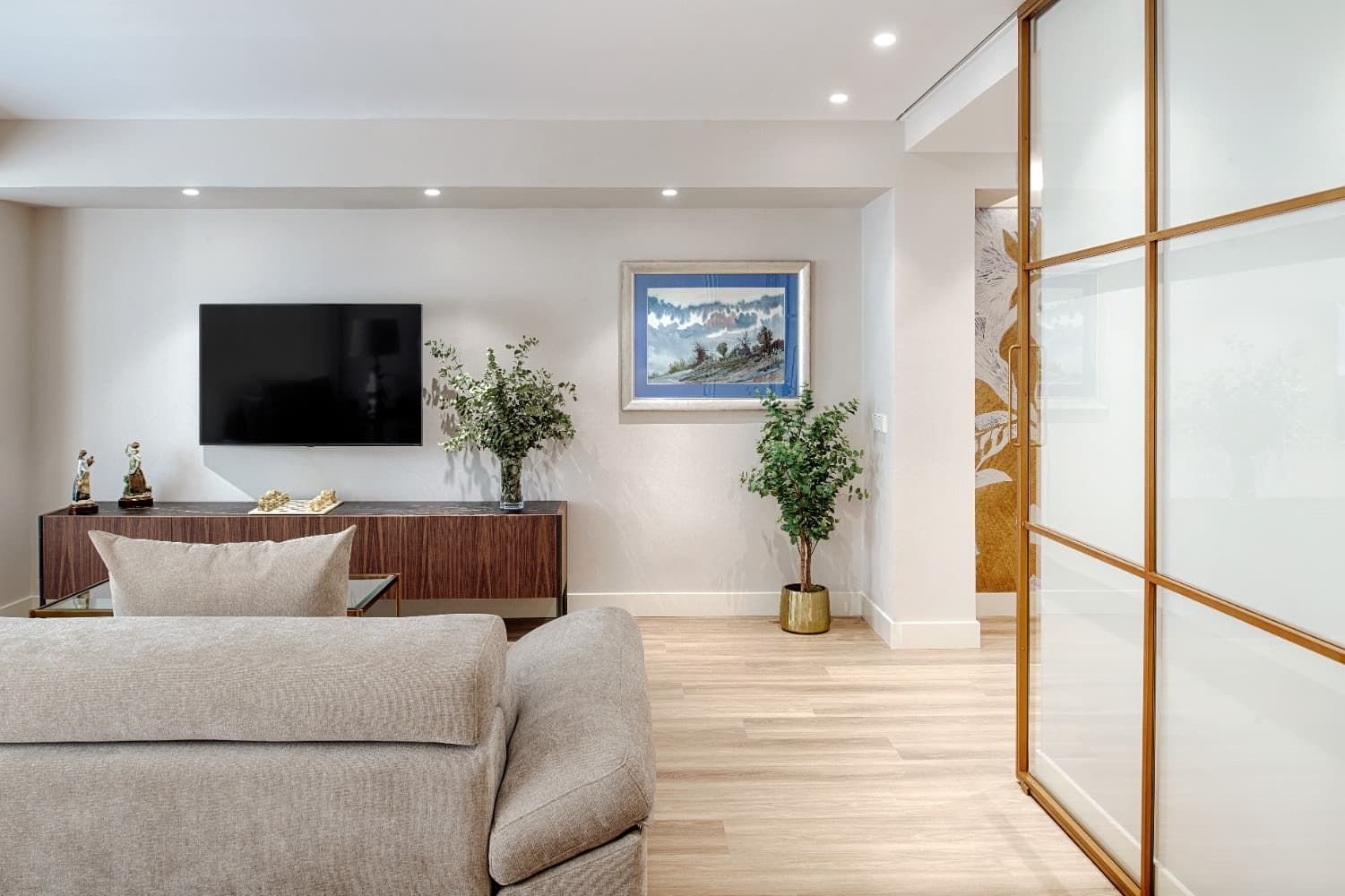 Sala de estar moderna con sofá gris, televisor montado en la pared, consola de madera, plantas y puerta corrediza de vidrio esmerilado.