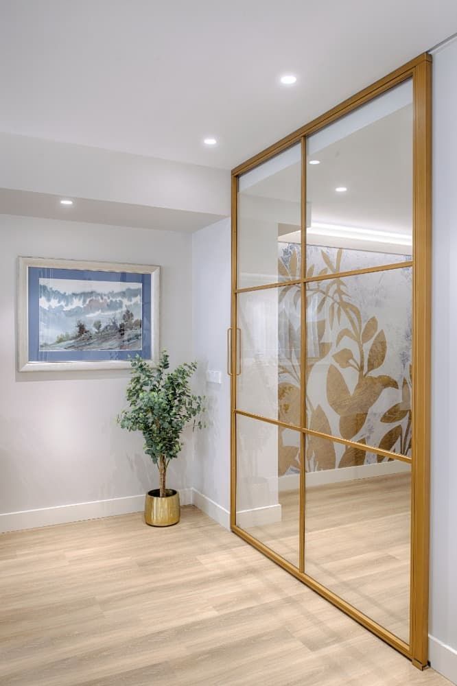 Pasillo moderno con puertas de cristal esmerilado con marcos dorados, suelo de madera clara, cuadros enmarcados y una planta en maceta.