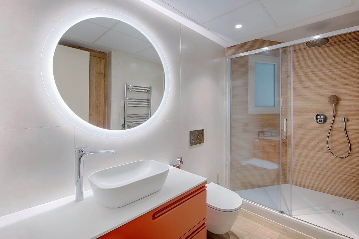 Baño moderno con espejo redondo retroiluminado, lavabo blanco, ducha y armarios de tocador naranjas.