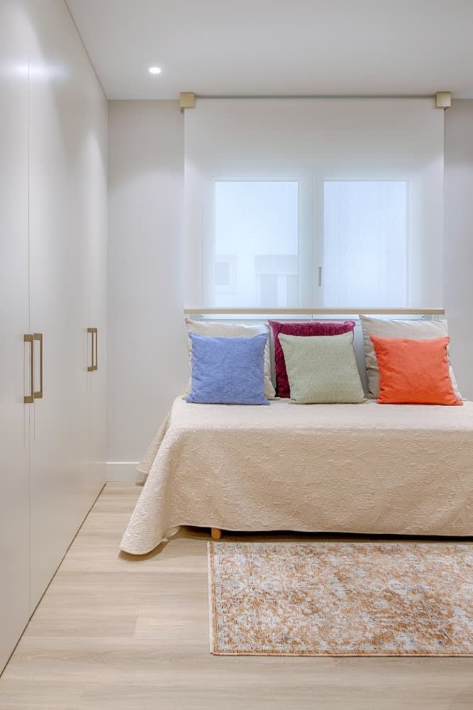 Dormitorio minimalista con cama, armario blanco y cojines de colores junto a una ventana luminosa.