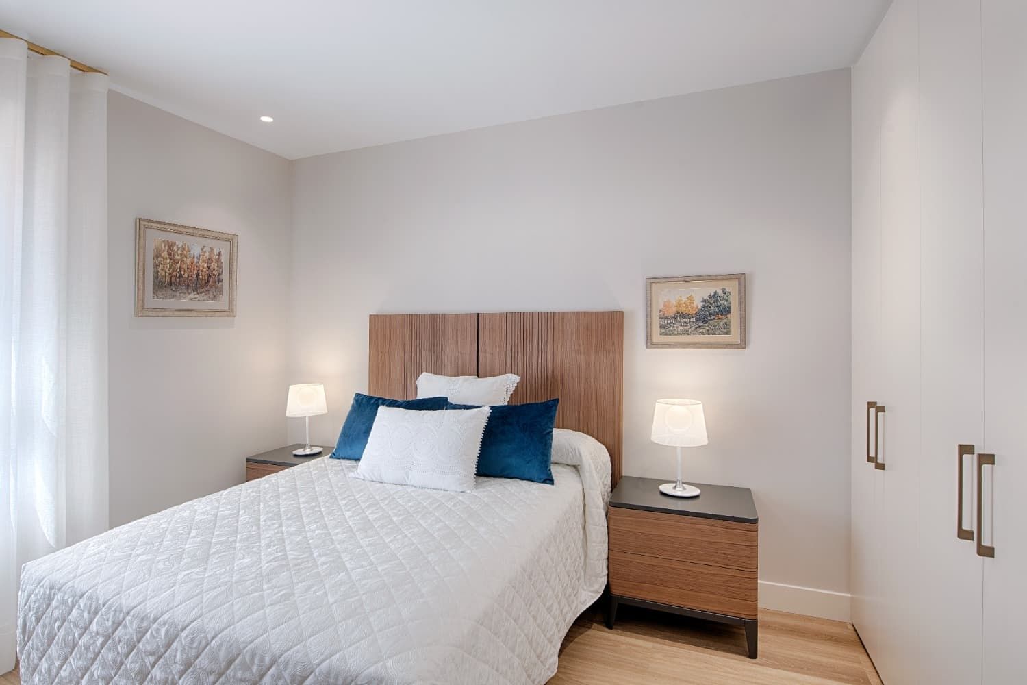 Habitación ordenada con ropa de cama blanca, mesitas de noche de madera, lámparas y cuadros enmarcados en paredes claras.