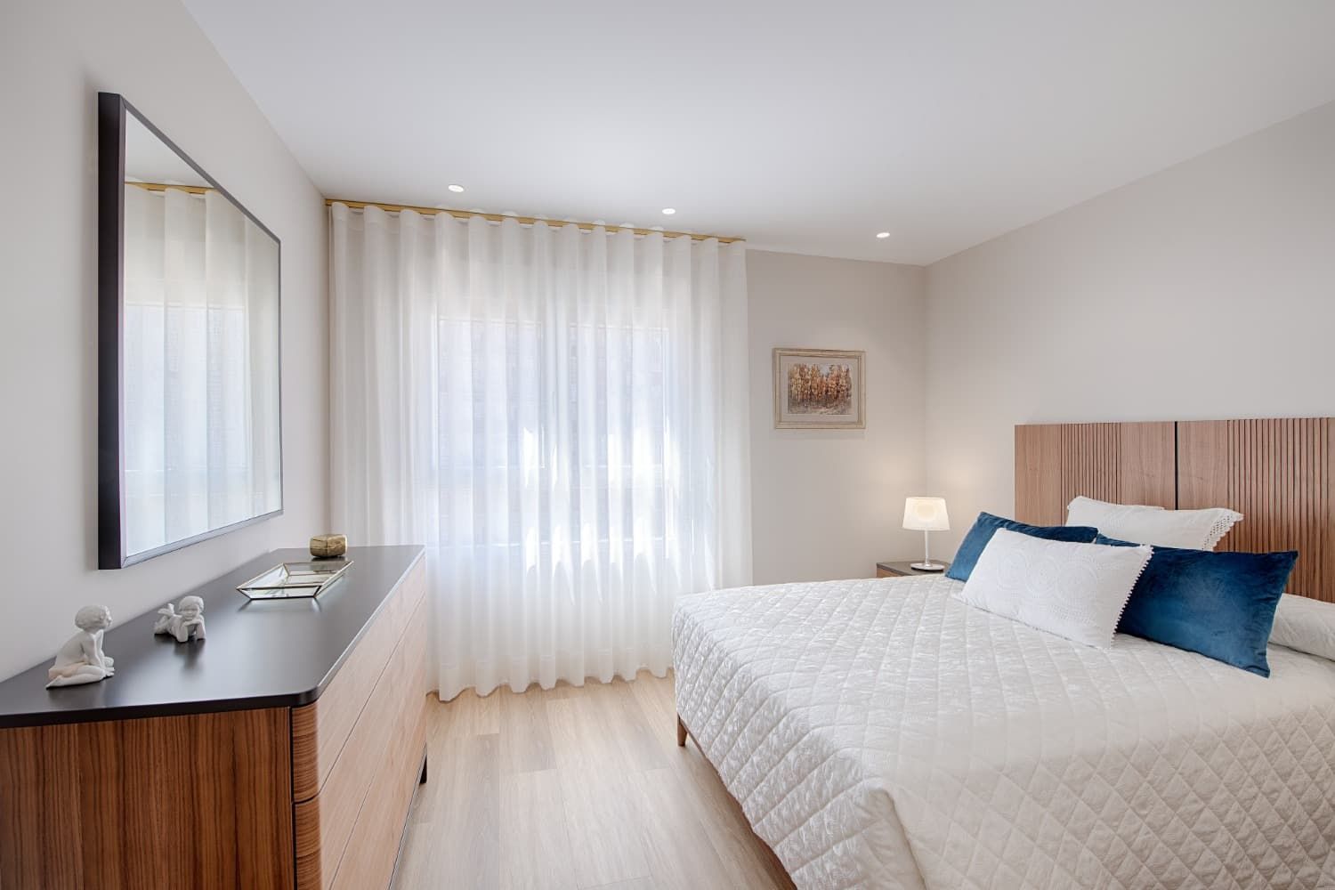 Habitación luminosa con cama blanca, cómoda de madera, cortinas transparentes y cuadros en tonos neutros suaves.