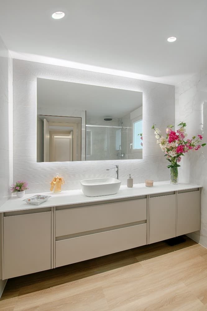 Tocador de baño moderno con un gran espejo retroiluminado, encimera blanca y flores rosas a la derecha.