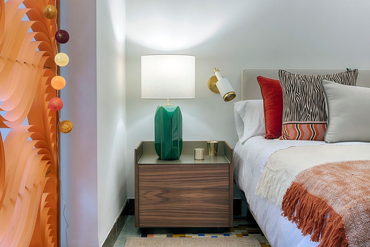 Un dormitorio con cama, mesita de noche, lámpara y cortina.