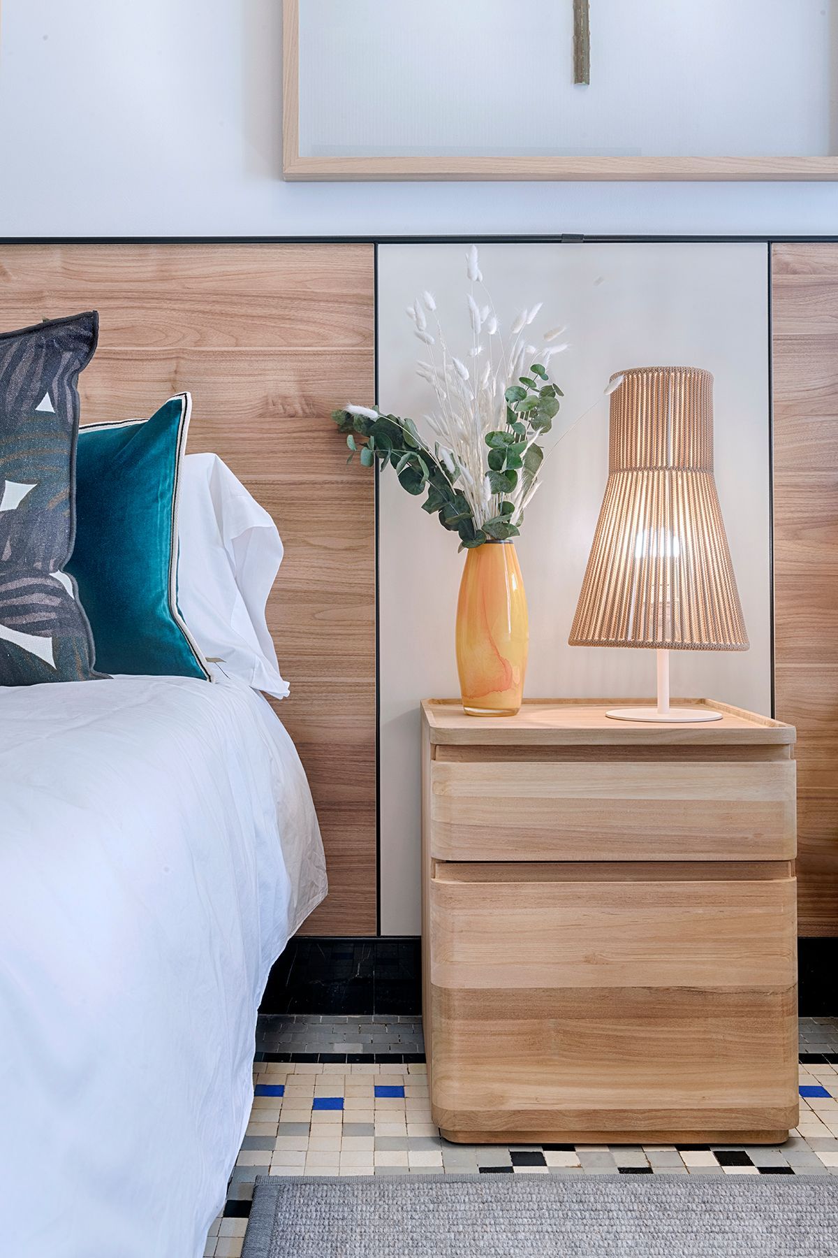 Un dormitorio con una cama, mesita de noche, lámpara y jarrón con flores.