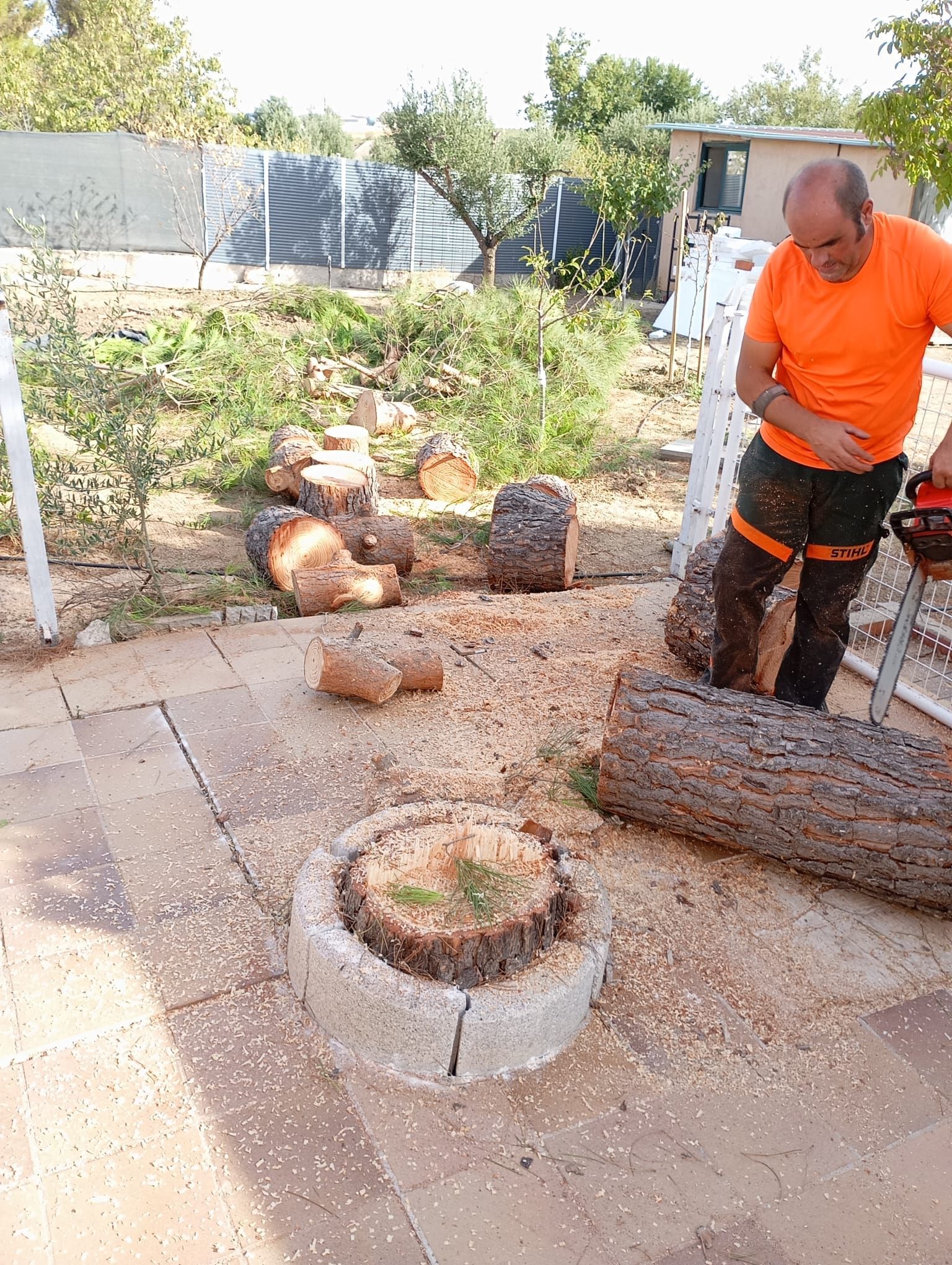 Hombre con camisa naranja cortando leña con una motosierra al aire libre. Trozos de madera y serrín en un patio de piedra.