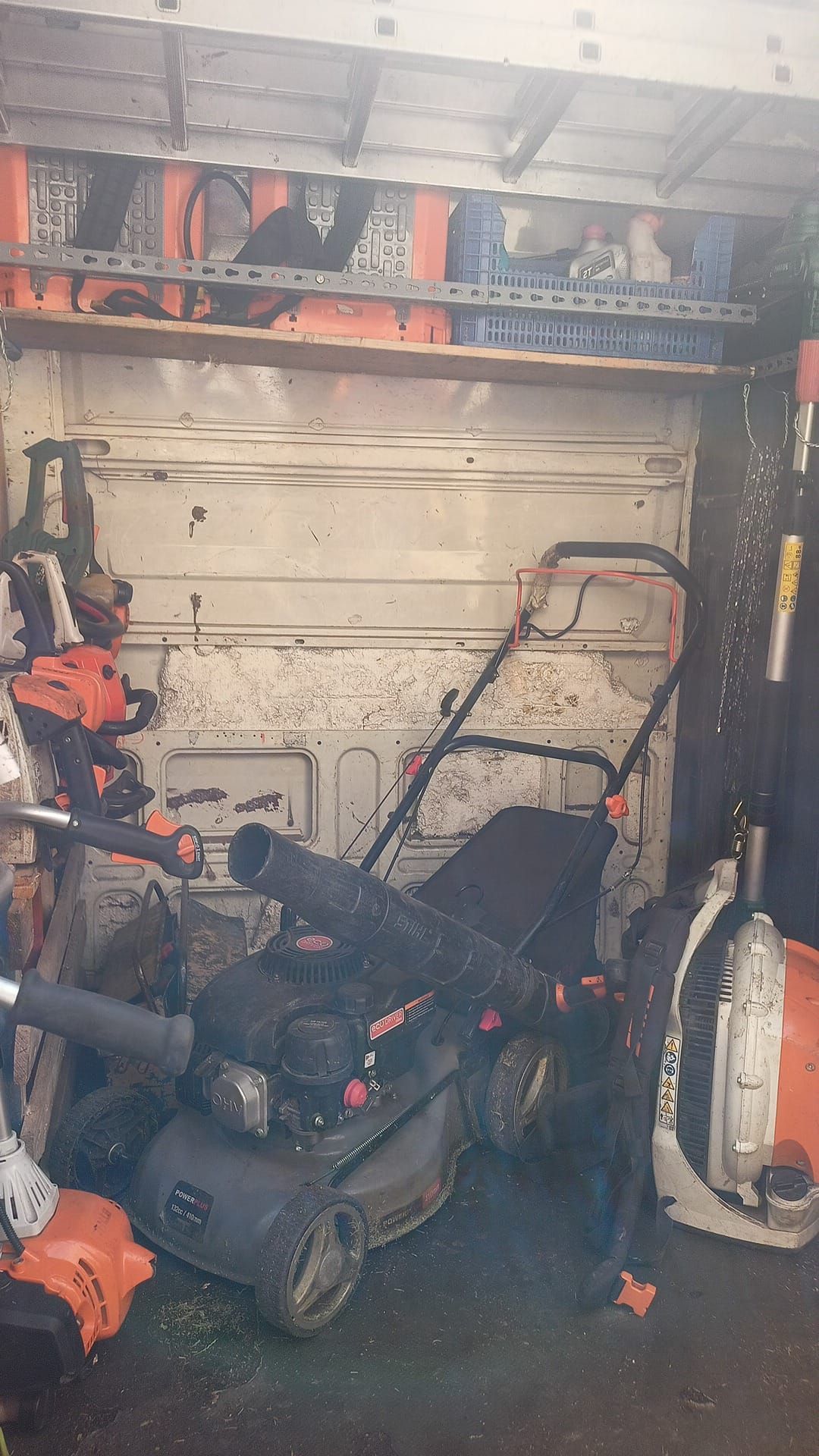 El interior de una furgoneta desordenada alberga equipos de jardinería: una cortadora de césped, un soplador y motosierras. Se ven herramientas naranjas y negras.