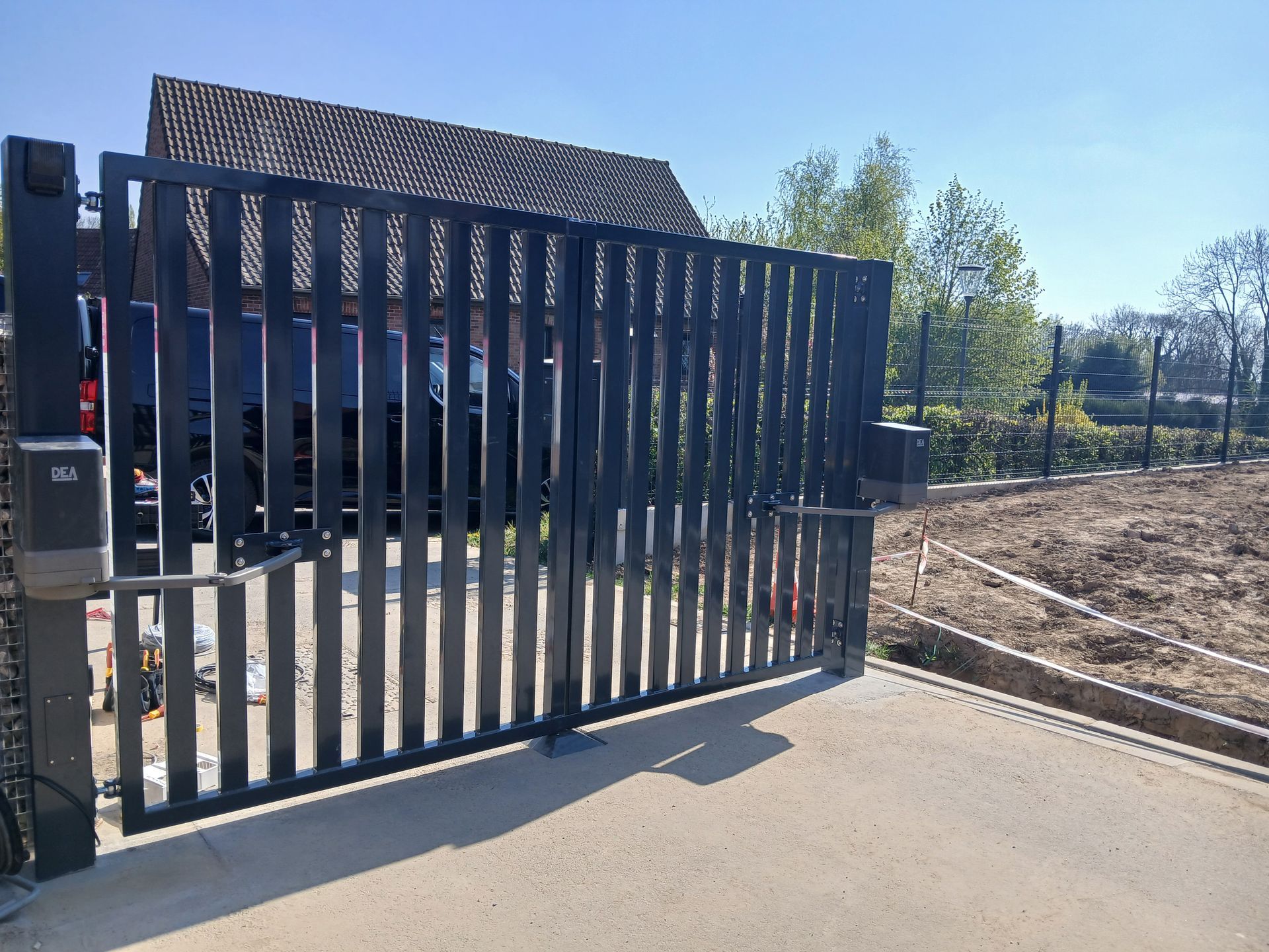 Portail noir à lattes verticales, ouvert sur une allée en béton, devant une maison.