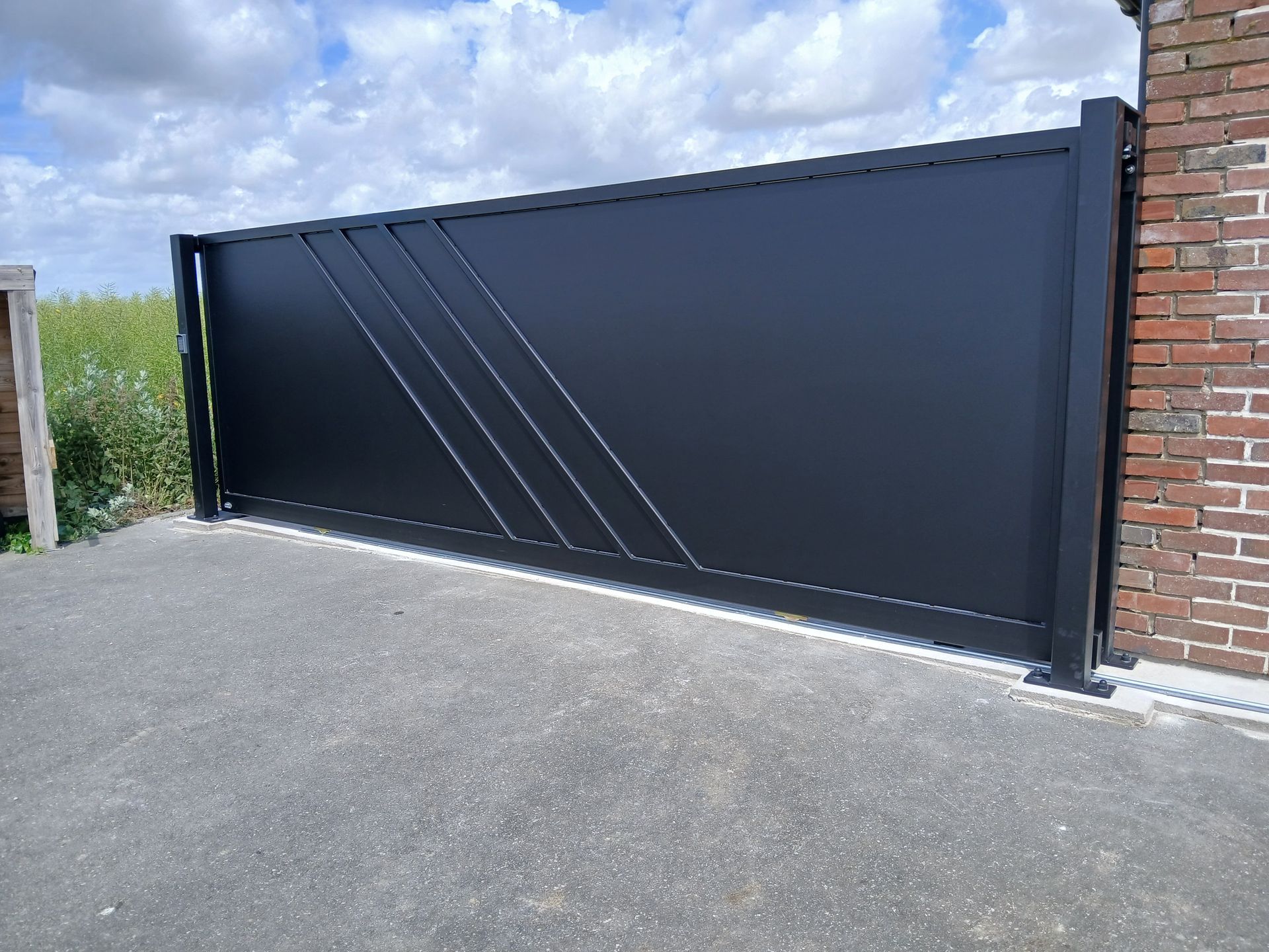 Portail coulissant noir avec design diagonal sur allée pavée, à côté d'un mur de briques.