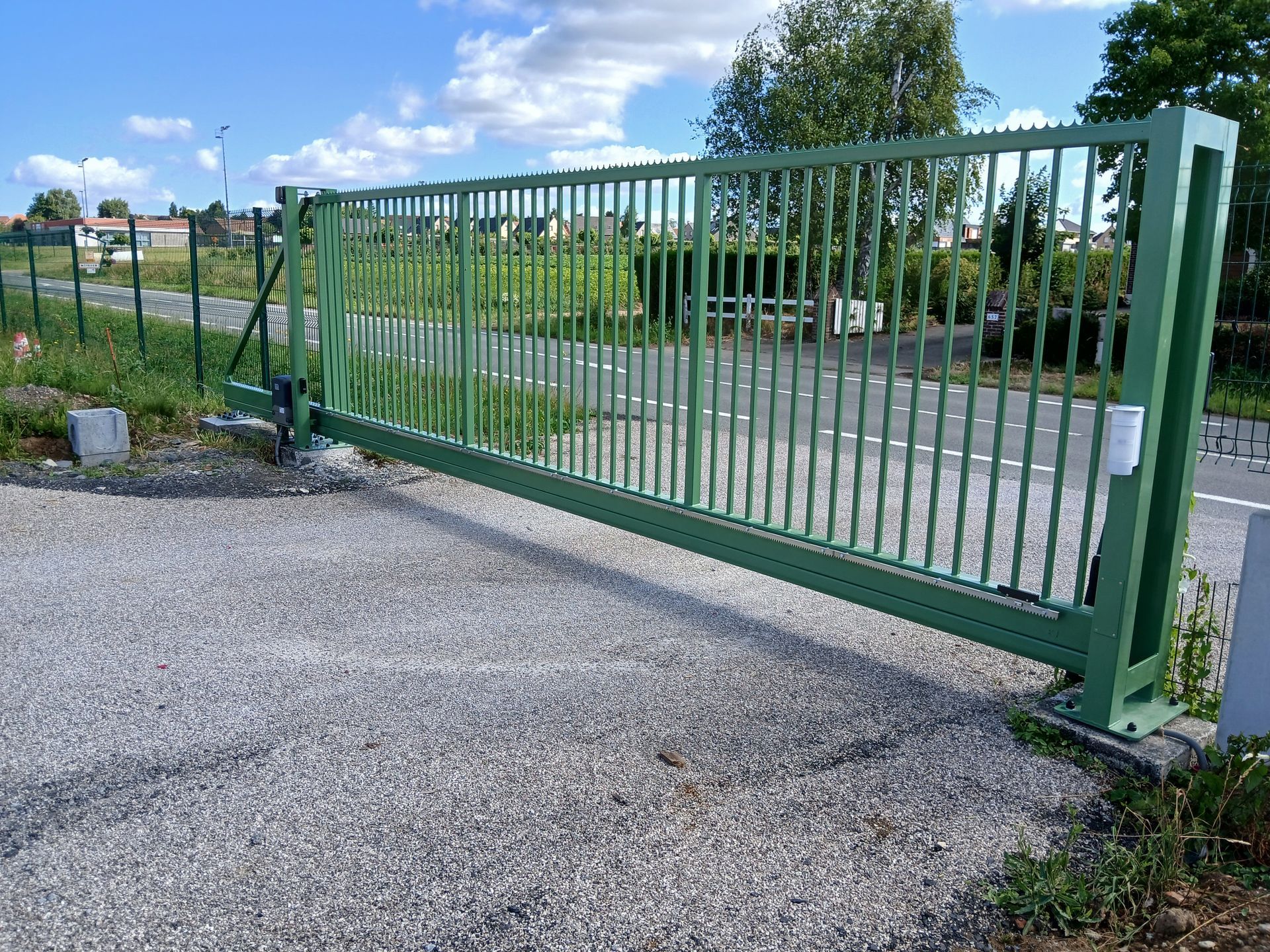 Portail coulissant en métal vert sur gravier, ouvert sur une route, avec verdure et bâtiments en arrière-plan.