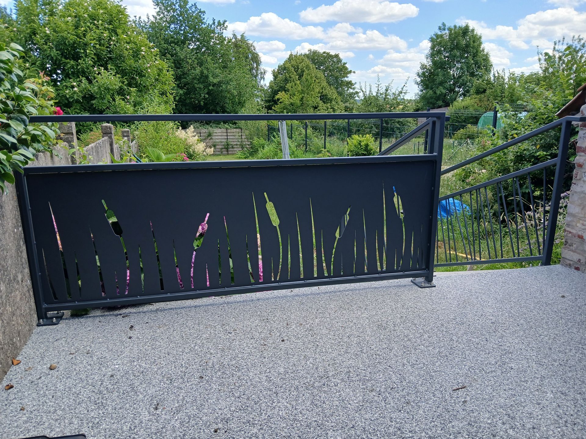 Portail en métal noir avec motif végétal découpé sur une terrasse en gravier.