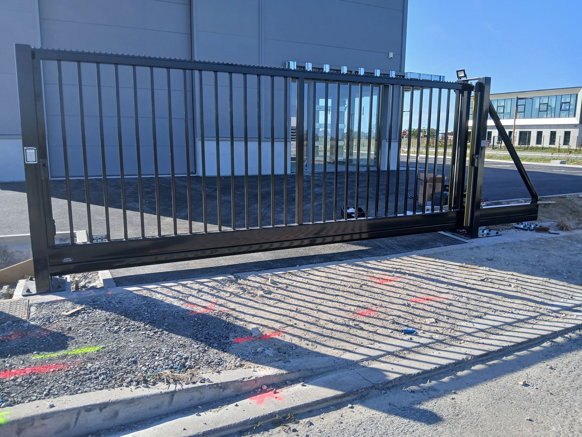 Portail coulissant en métal noir devant un immeuble. Surface en béton et gravier.
