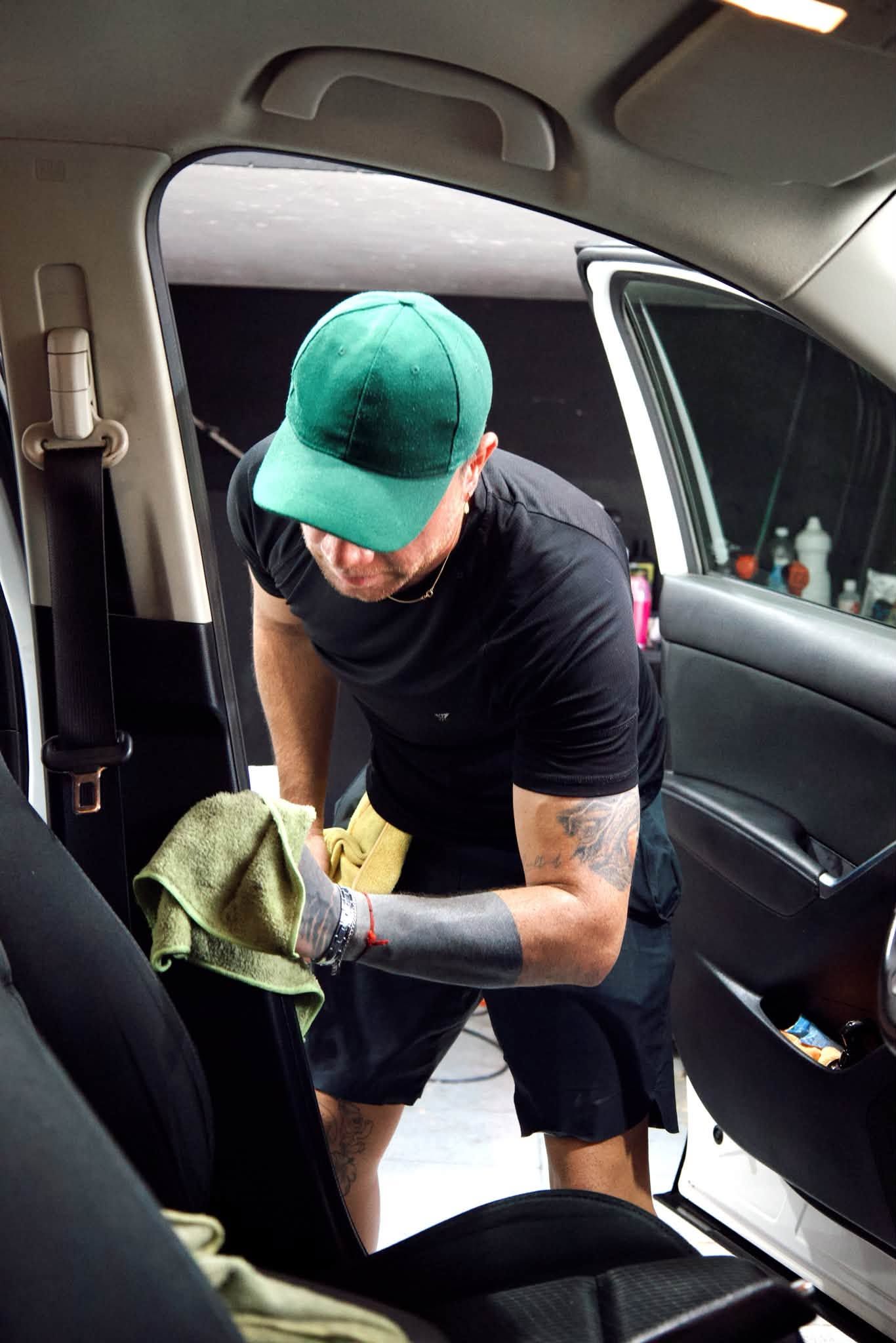 Una persona limpiando el interior de un coche con un paño, con una gorra verde y una camiseta negra.