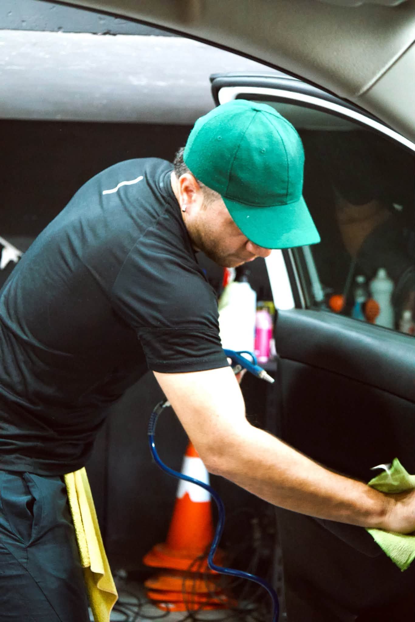 Un hombre con una gorra verde limpia el interior de un coche con un paño, junto a la puerta abierta