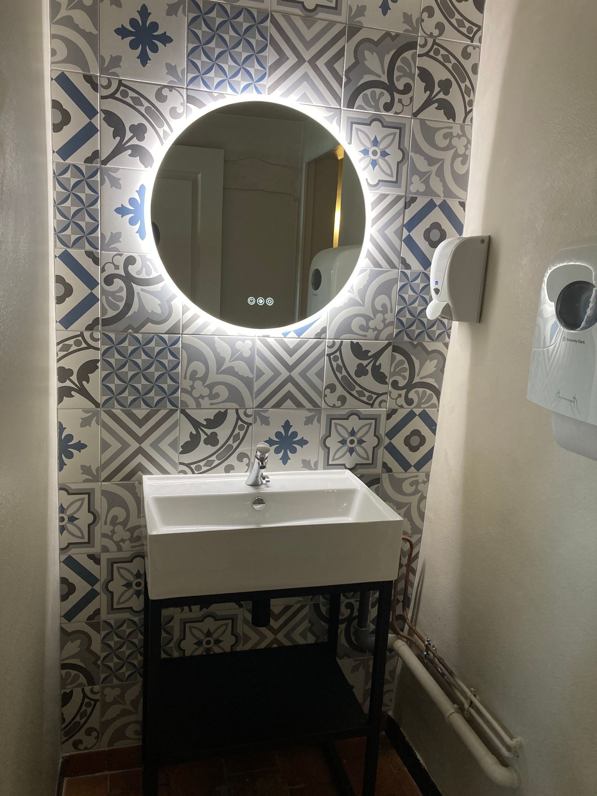 Petit lavabo de salle de bains avec miroir rond rétroéclairé et mur carrelé de carreaux bleus et blancs à motifs.