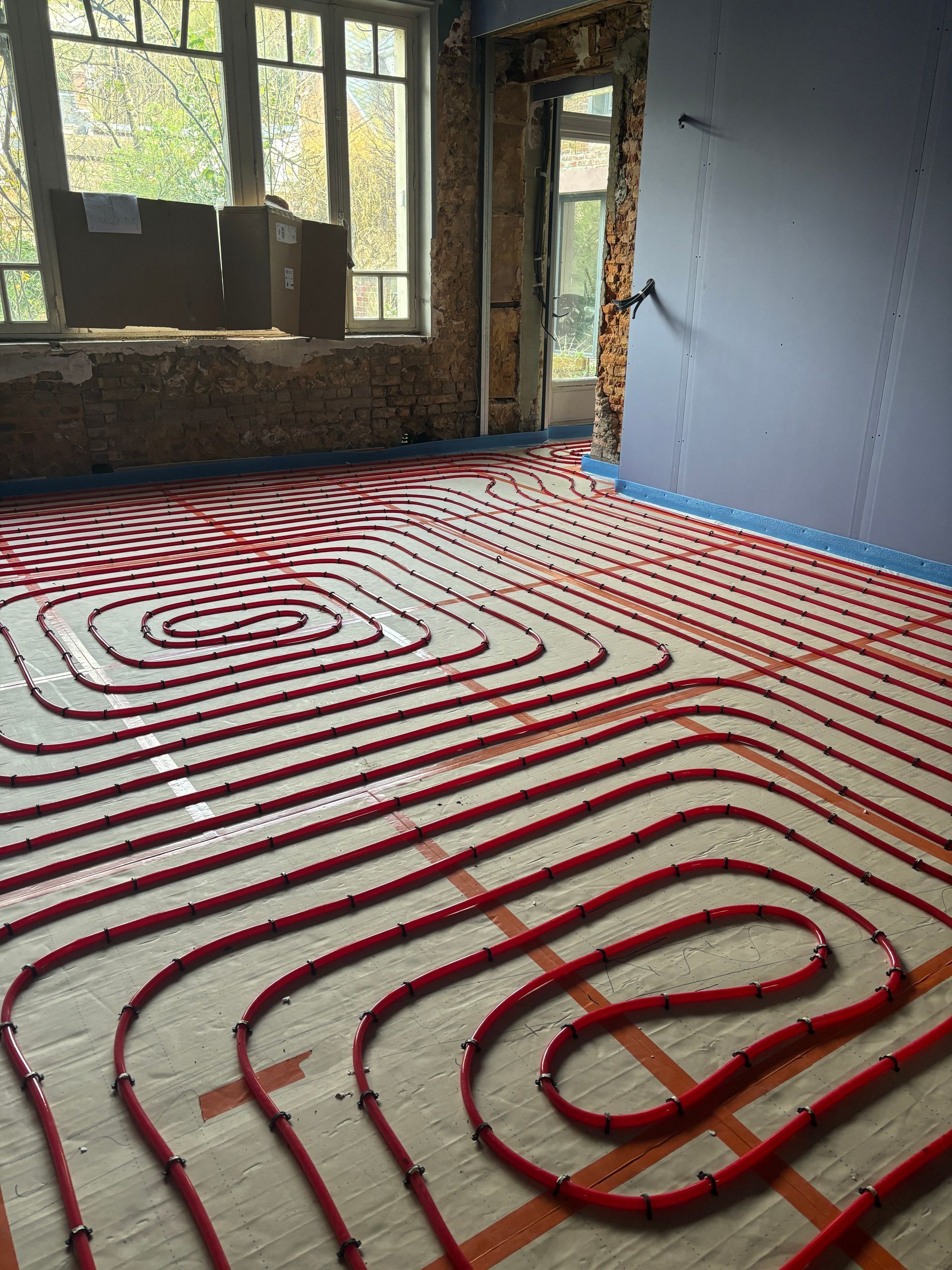 Des tuyaux de chauffage au sol rouges sont installés dans une pièce en construction avec des fenêtres et des murs non finis.