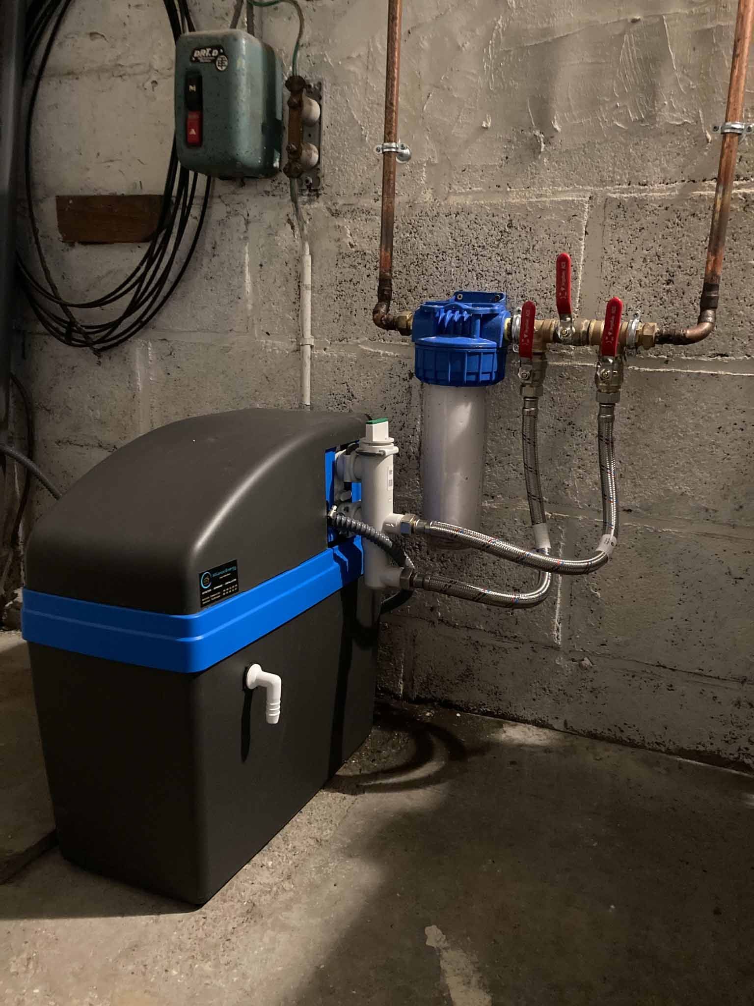 Système de traitement des eaux avec réservoir noir, boîtier de filtre bleu et commandes murales dans un sous-sol.
