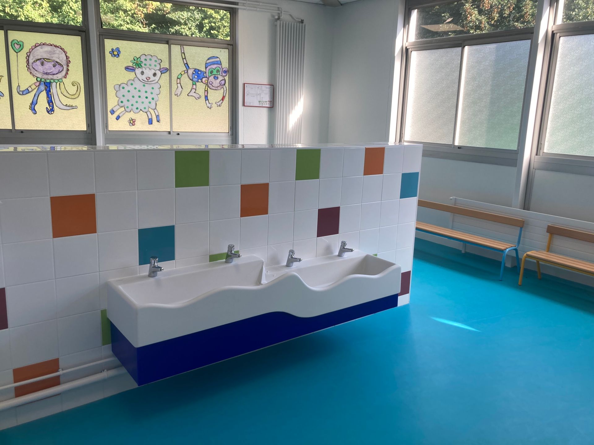 Salle de bains lumineuse de garderie avec lavabos blancs, sol bleu, mur carrelé et dessins d'enfants sur les fenêtres.