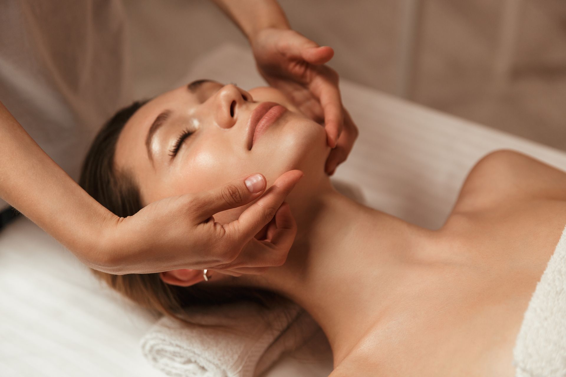 Des mains massent délicatement le visage d'une femme dans un spa, enveloppée dans une serviette blanche et baignée d'une douce lumière beige.