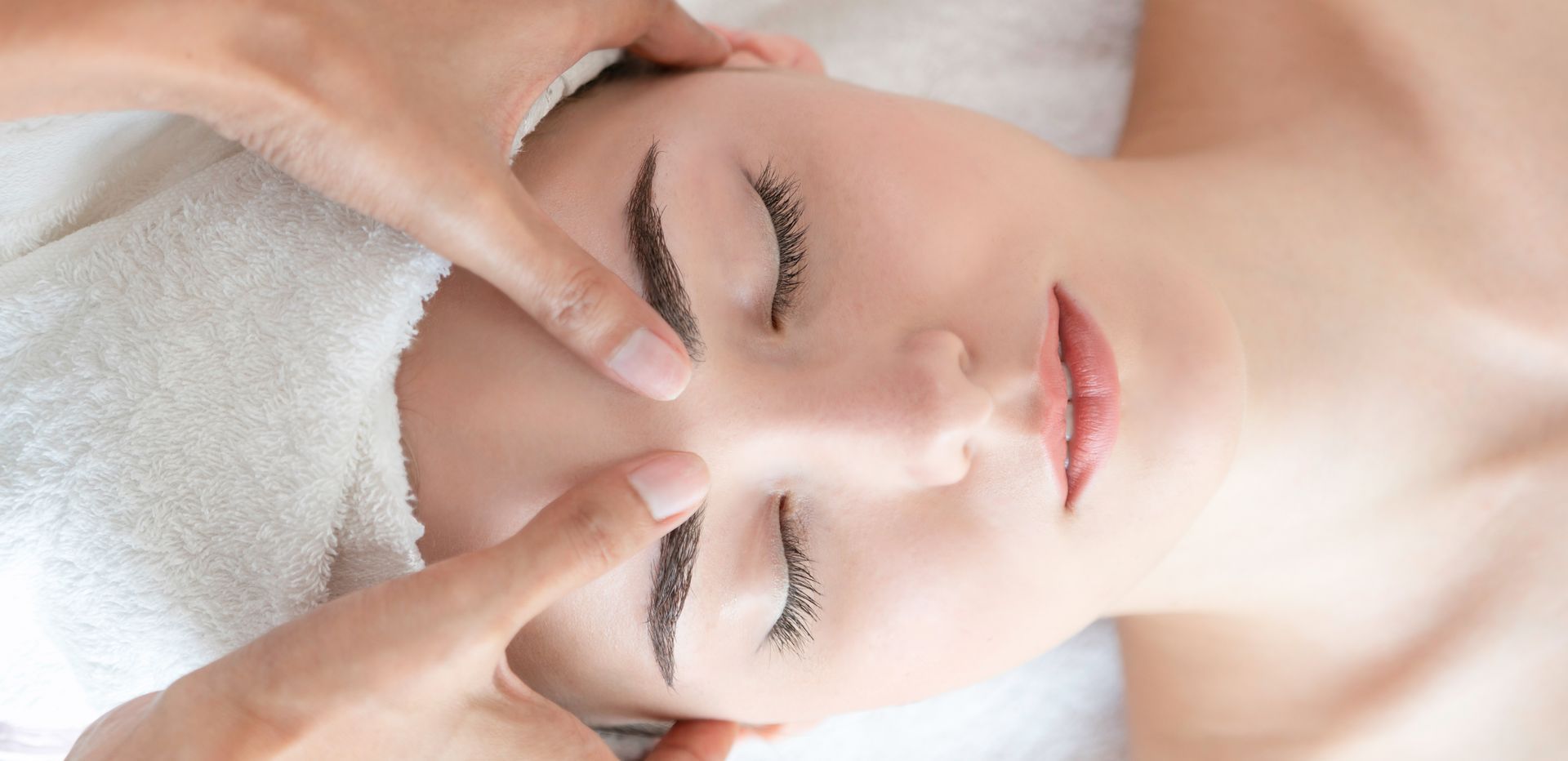 Gros plan sur le visage d'une femme aux yeux fermés recevant un massage facial sur une serviette blanche.