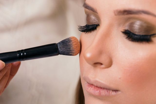 Un pinceau de maquillage applique du blush sur la joue d'une femme aux yeux fermés.