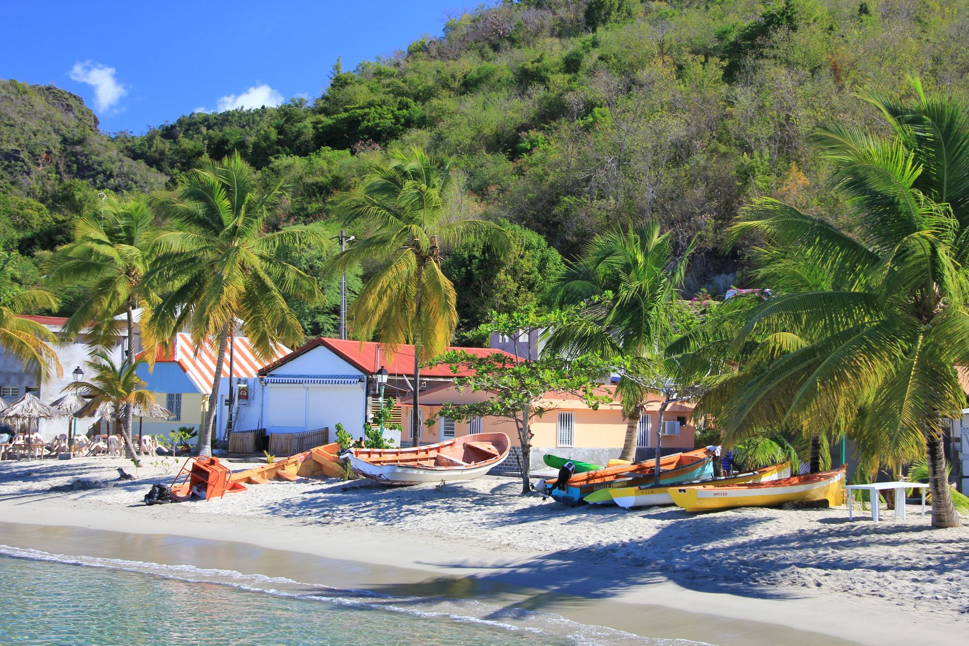 Plage tropicale avec palmiers, bateaux colorés et petites maisons à flanc de colline verdoyante.