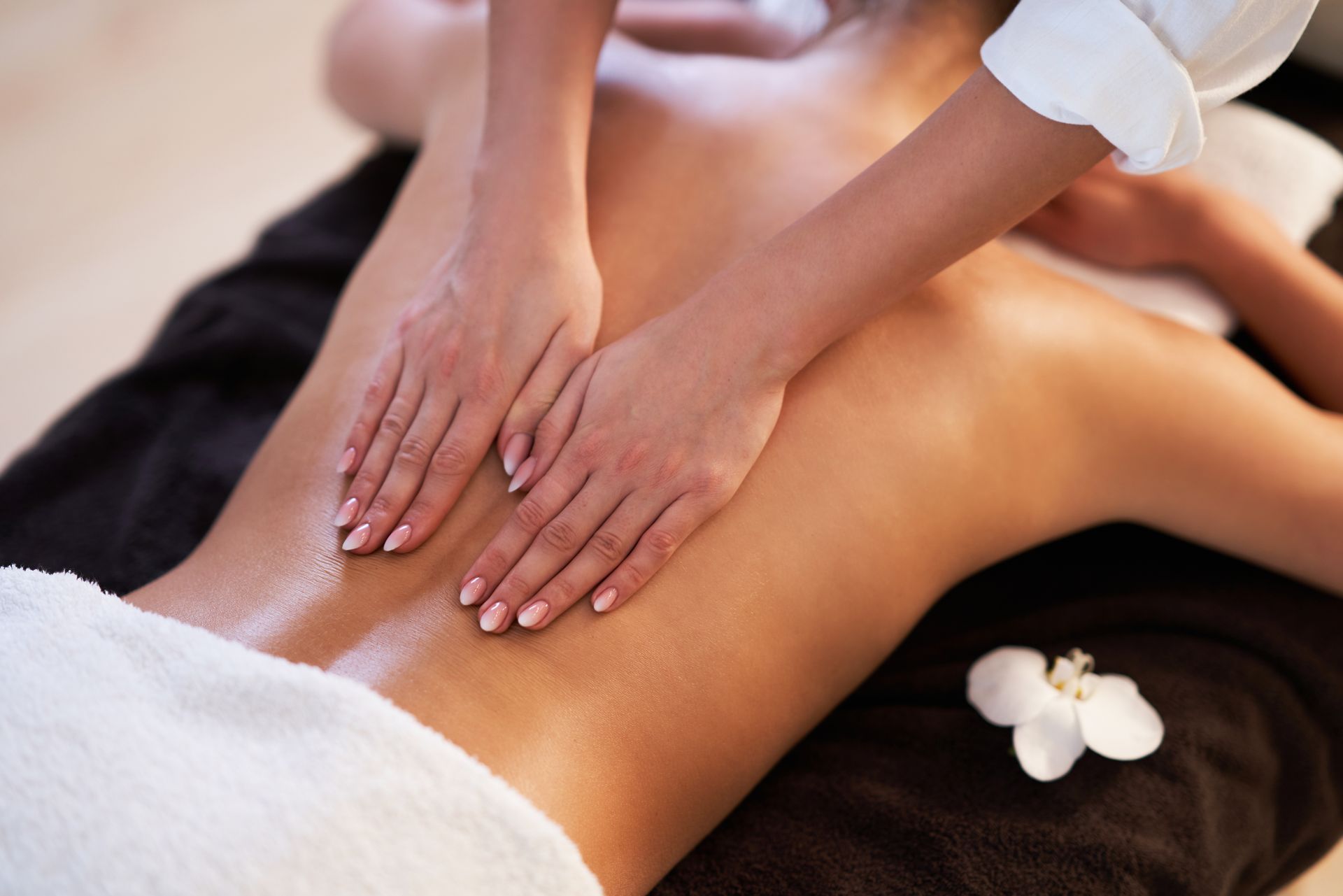 Une masseuse applique de l'huile sur le dos d'une personne, avec des touches de fleurs blanches.