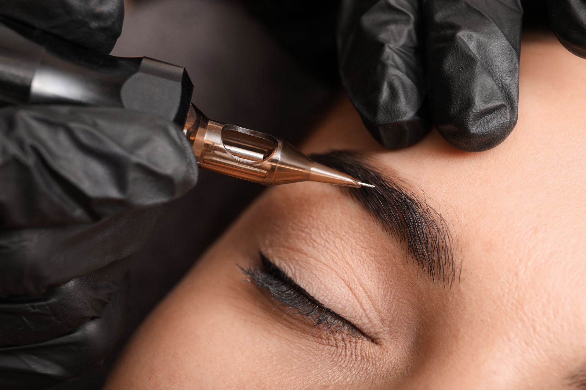 Des mains gantées appliquent une teinture pour sourcils au pinceau sur un œil fermé lors d'un soin esthétique.