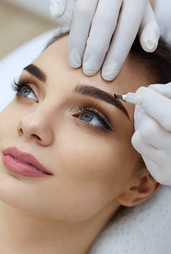 Des mains gantées sculptent les sourcils d'une femme allongée, le regard tourné vers le ciel, pendant un soin de beauté.