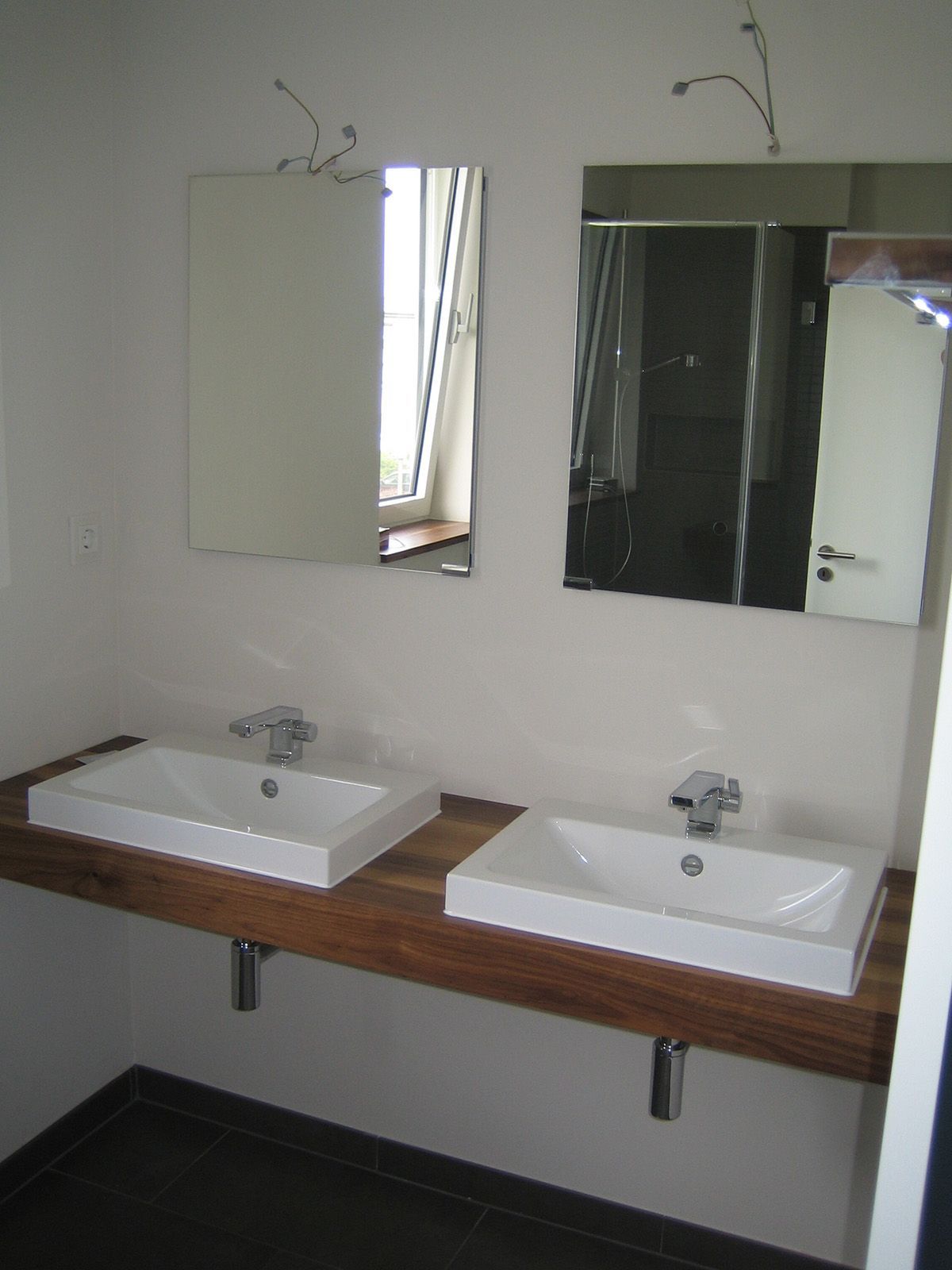 Zwei weiße Waschbecken auf einer Holztheke, darüber Spiegel. Chromhähne, dunkler Boden und weiße Wände.