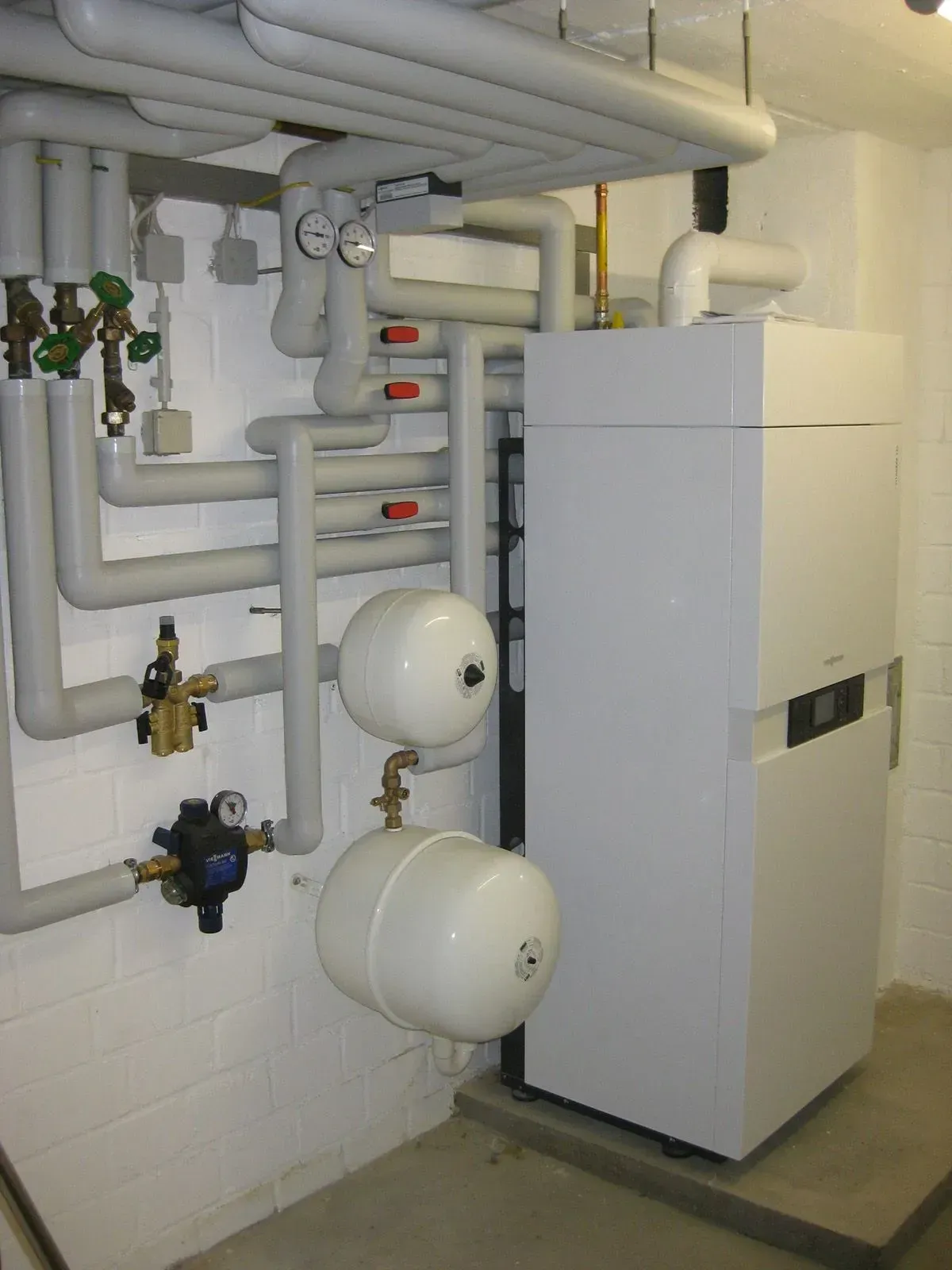 Heizsystem mit Rohren, Tanks und einer weißen Einheit in einem Keller.