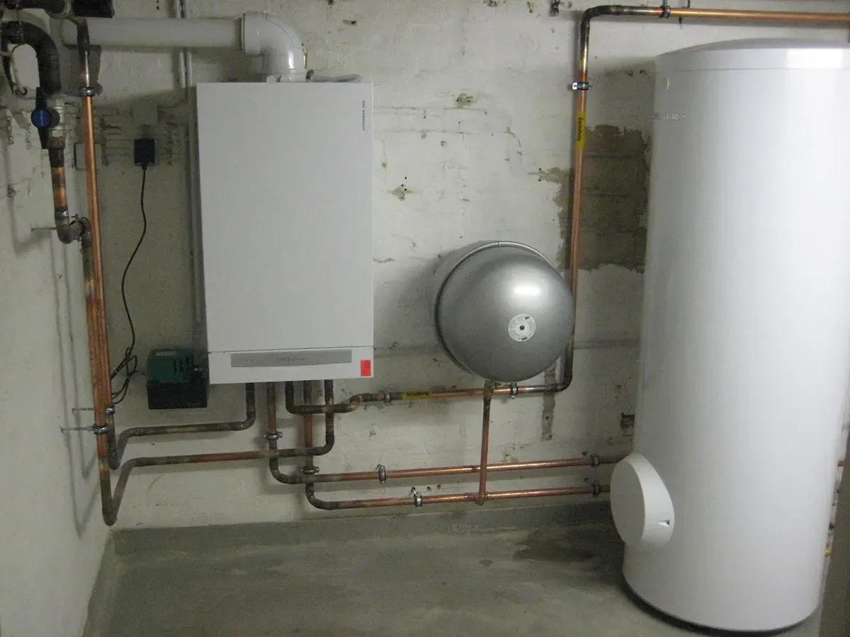 Weißes Warmwasserbereitersystem mit Kupferrohren, einem Druckbehälter und einem Boiler in einem Hauswirtschaftsraum.