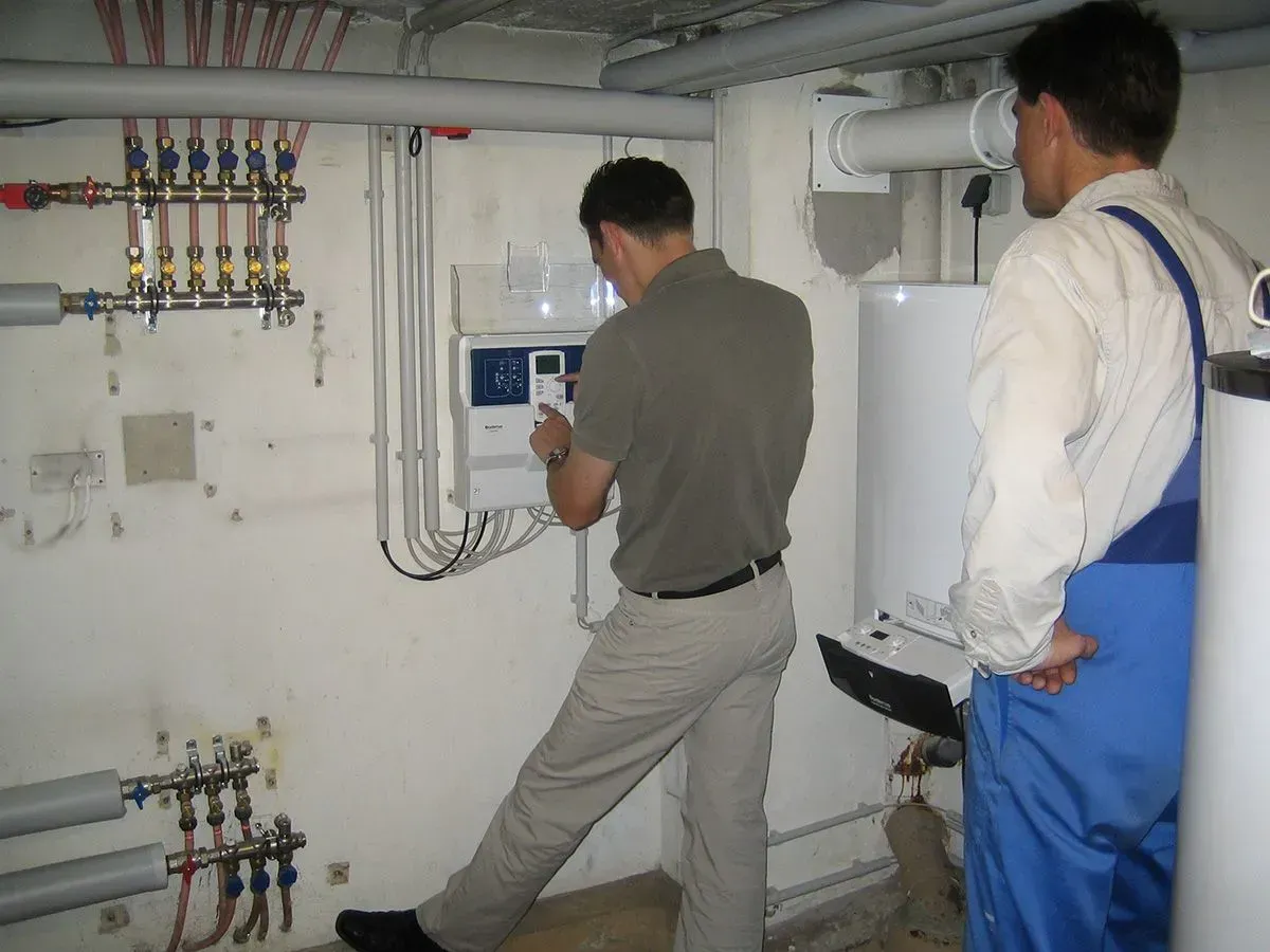 Zwei Männer untersuchen die Steuerung einer Heizung in einem Hauswirtschaftsraum. Einer zeigt, der andere beobachtet.