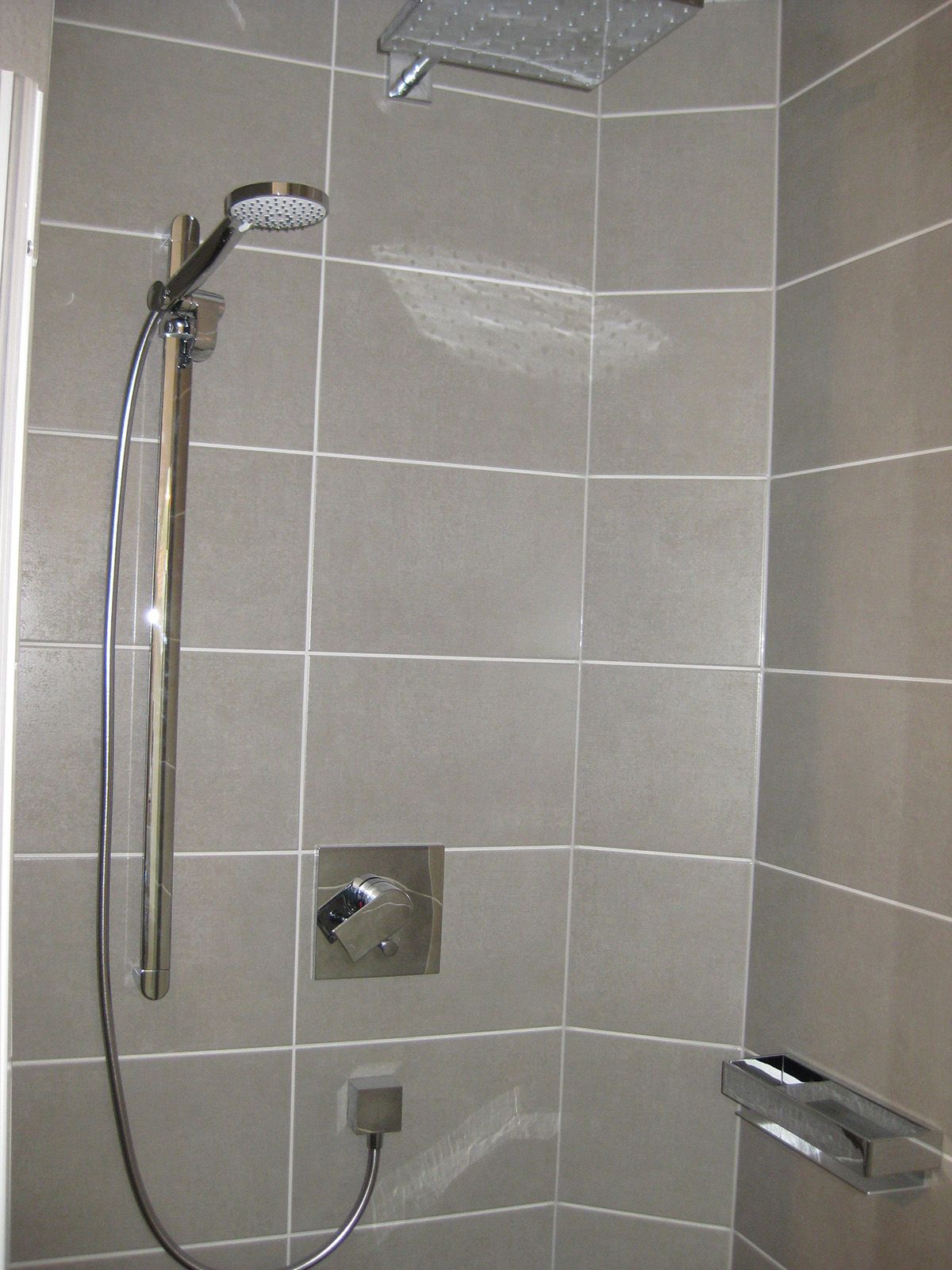 Grau geflieste Dusche mit Chromarmaturen: Handbrause, Regendusche und Bedienknopf.