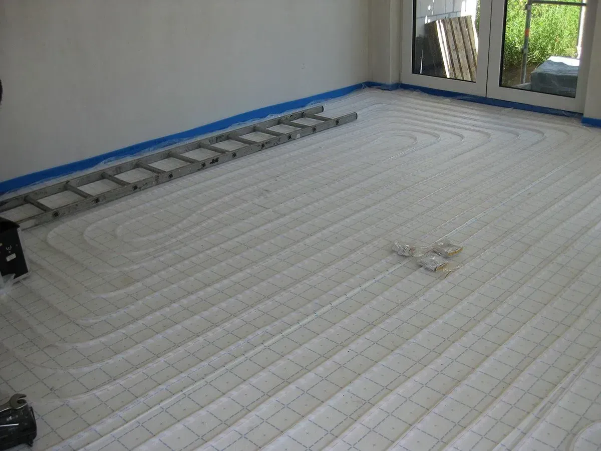 Weißer Boden mit eingelassenen Rohren für die Strahlungsheizung, Leiter im Hintergrund, blaues Klebeband entlang der Wand.