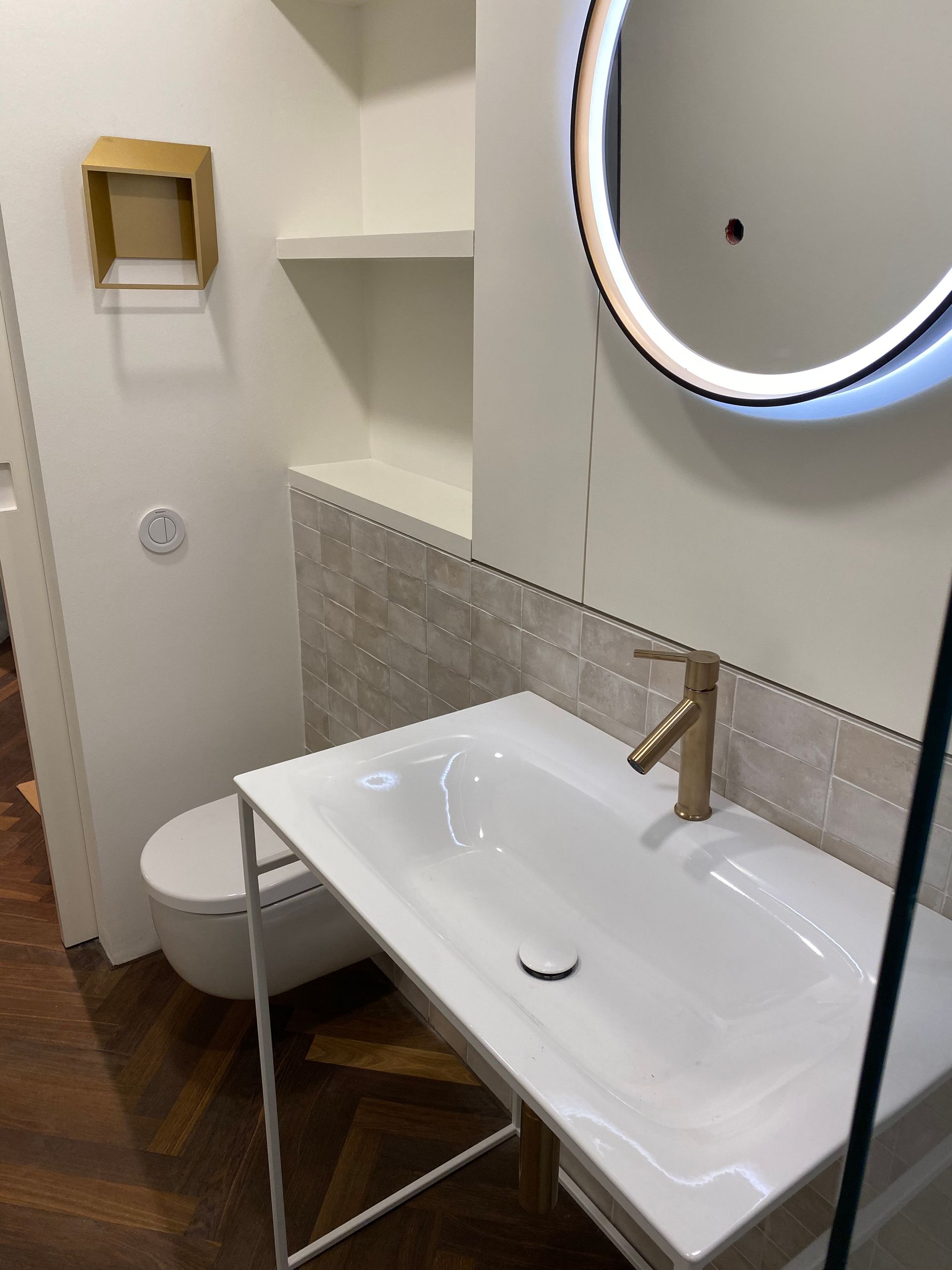 Modernes Badezimmer mit weißem Waschbecken, goldener Armatur, rundem Spiegel und Toilette.