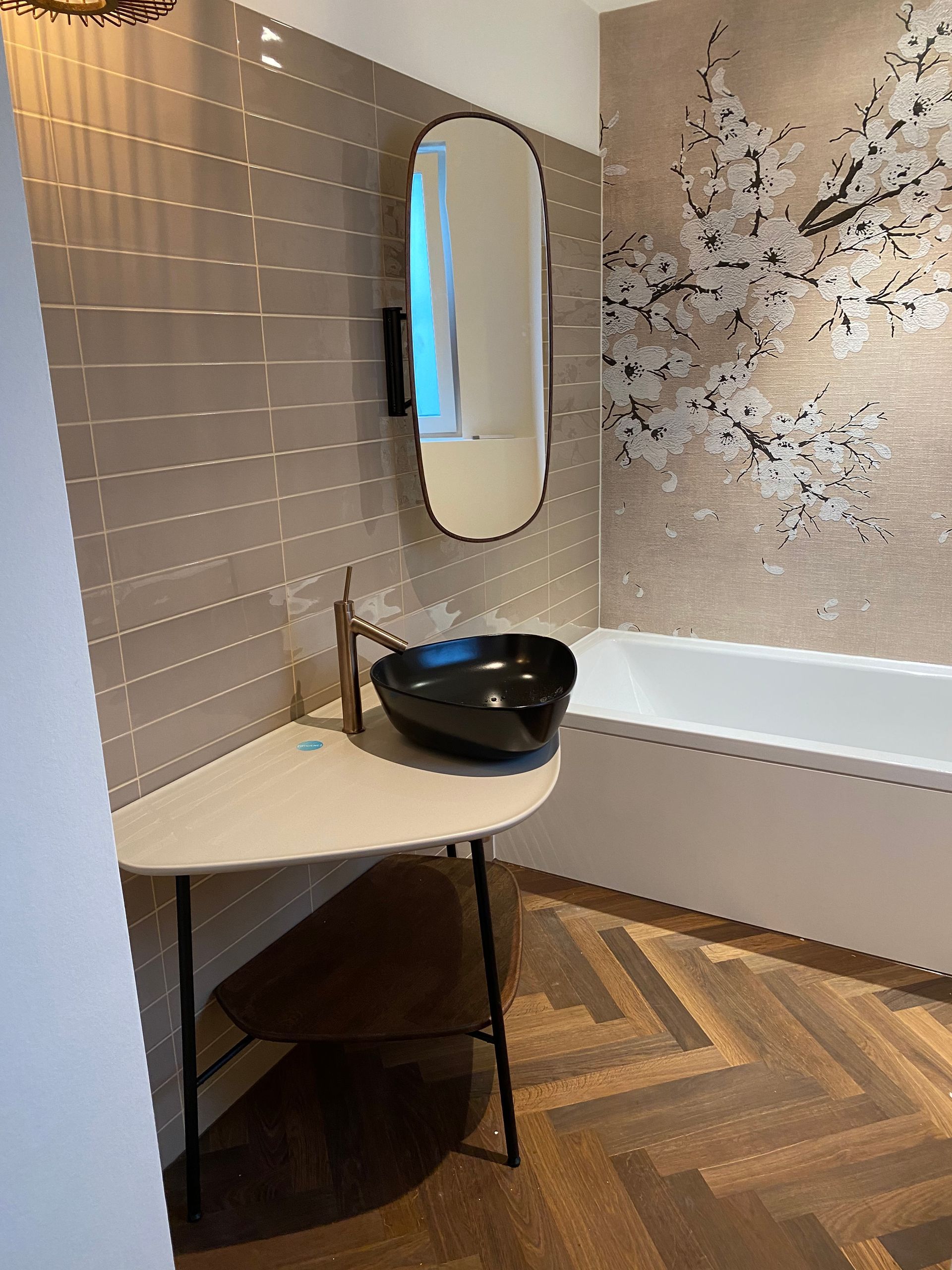 Badezimmer mit schwarzem Waschbecken, ovalem Spiegel, gemusterter Wand und Holzboden.