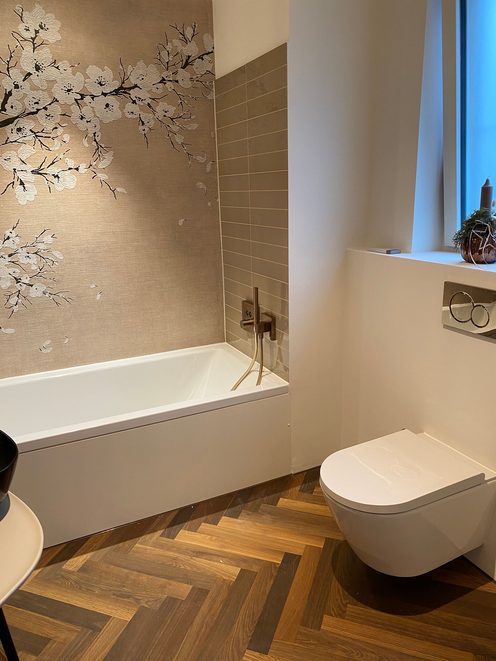 Badezimmer mit weißer Badewanne und Toilette, braunen und beigen Fliesen, Holzboden und floraler Wandkunst.