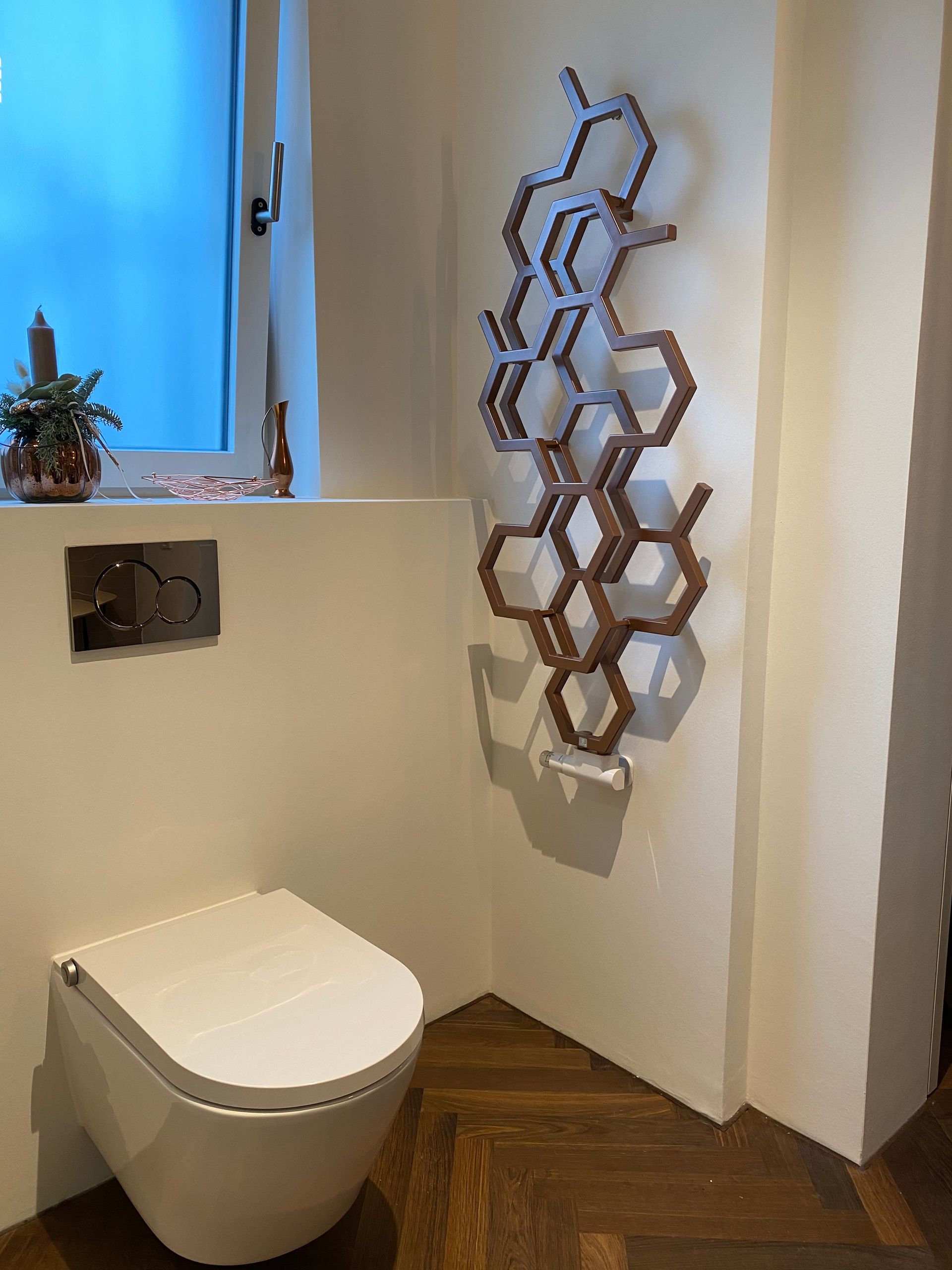 Ein modernes Badezimmer mit weißer Toilette, wabenförmigem Heizkörper und kleinem Fenster; brauner Parkettboden.