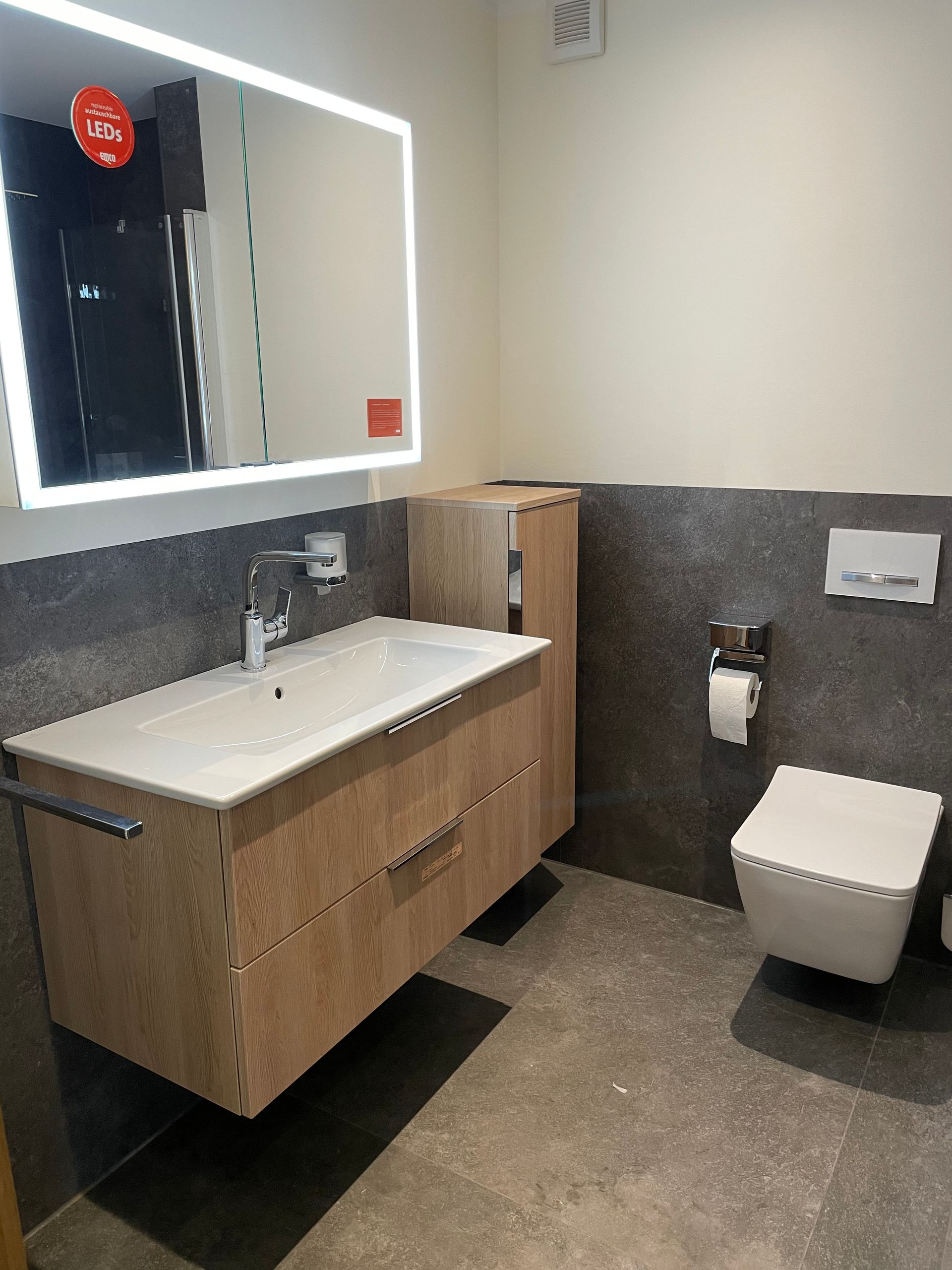 Modernes Badezimmer mit schwebendem Waschtisch aus Holz, quadratischer Toilette und beleuchtetem Spiegel.