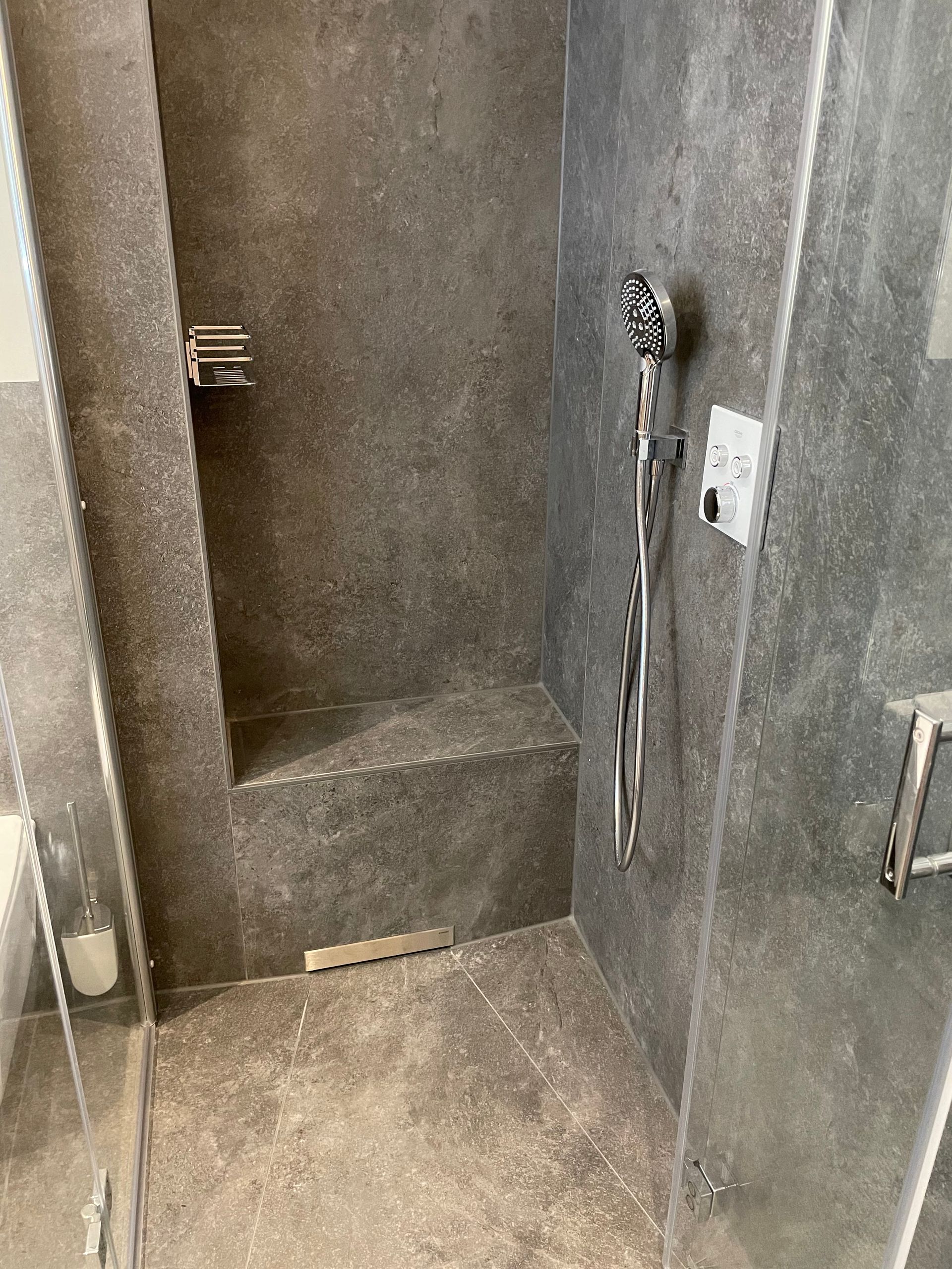 Grau geflieste Dusche mit integriertem Sitz, Glastür und silbernem Duschkopf.