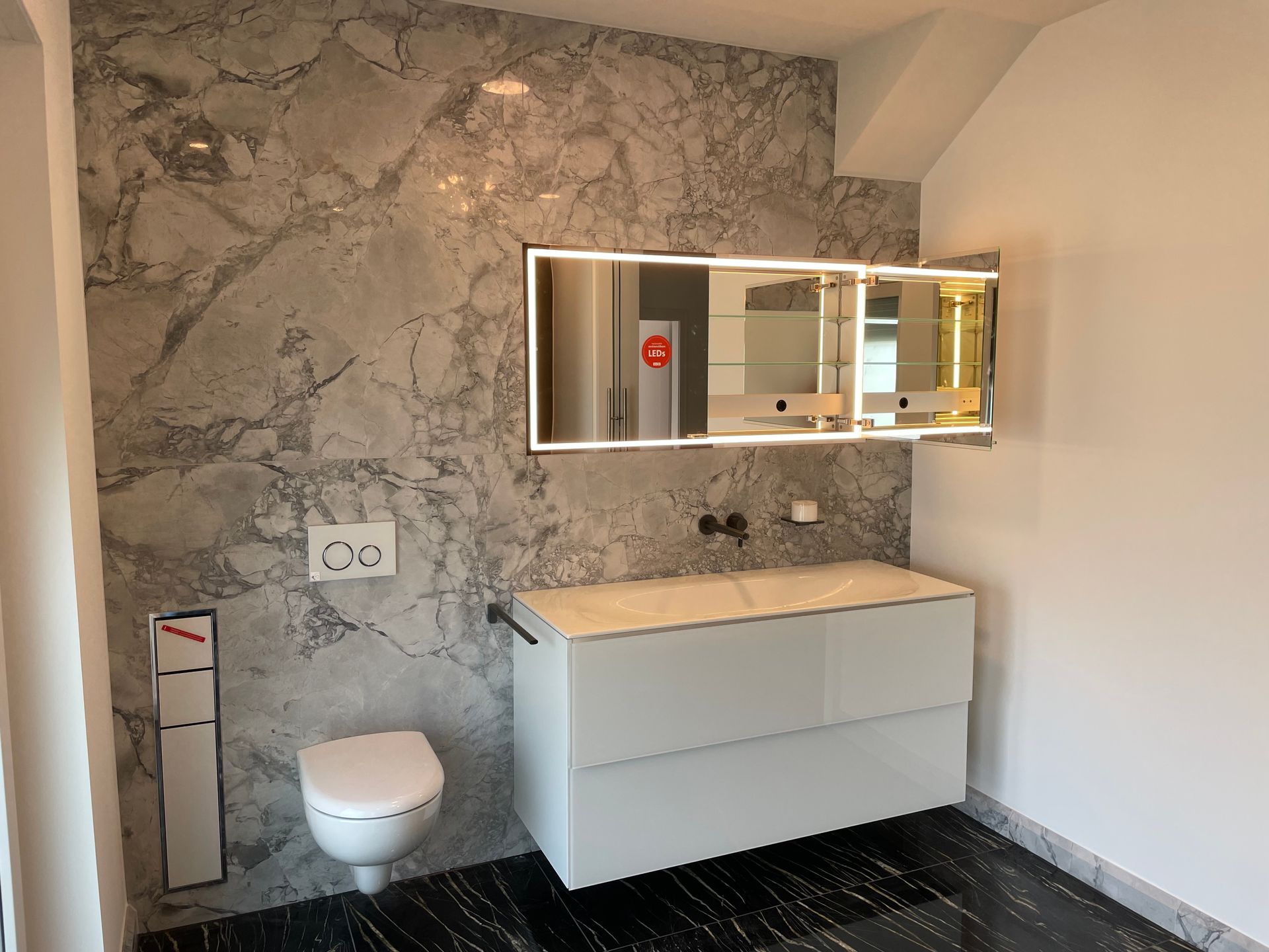 Badezimmer mit Marmorwand, weißem Waschtisch, Toilette und beleuchtetem Spiegel. Schwarzer Boden.