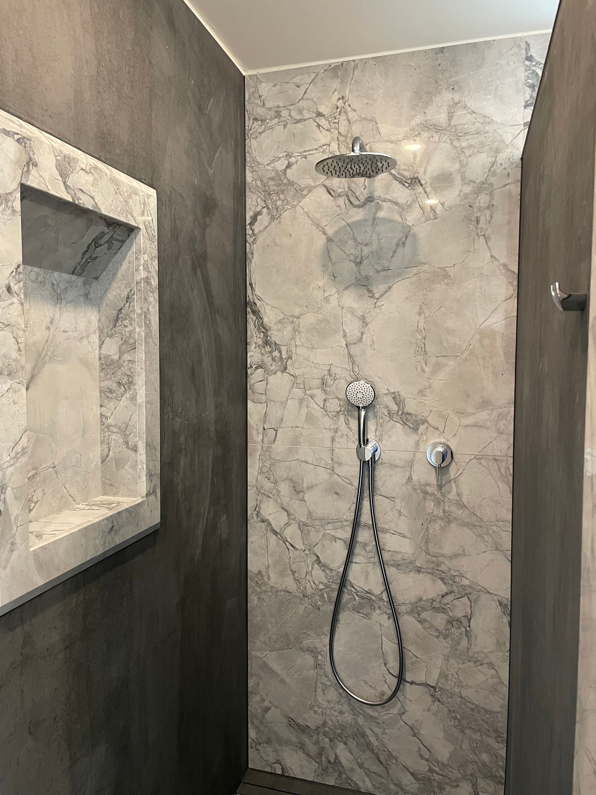 Dusche mit Marmorwand und integriertem Regal. Dunkelgraue Wände. Verchromter Duschkopf und Handbrause.