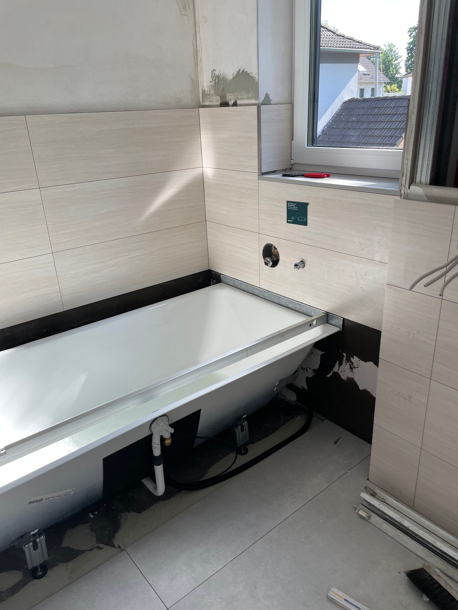 Badezimmer im Bau; weiße Badewanne, geflieste Wände, Fenster, sichtbare Rohrleitungen.