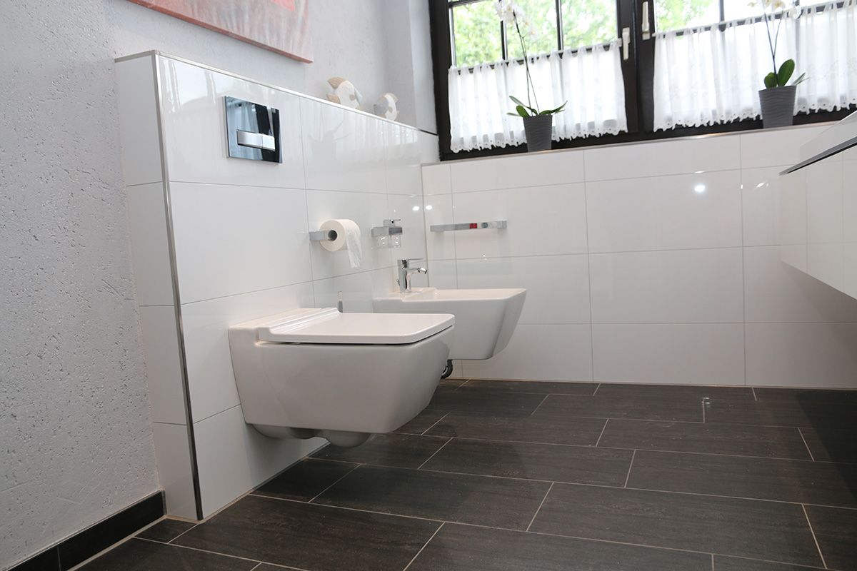 Modernes weißes Badezimmer mit wandmontiertem WC und Bidet, dunklem Fliesenboden und einem Fenster mit weißen Vorhängen.