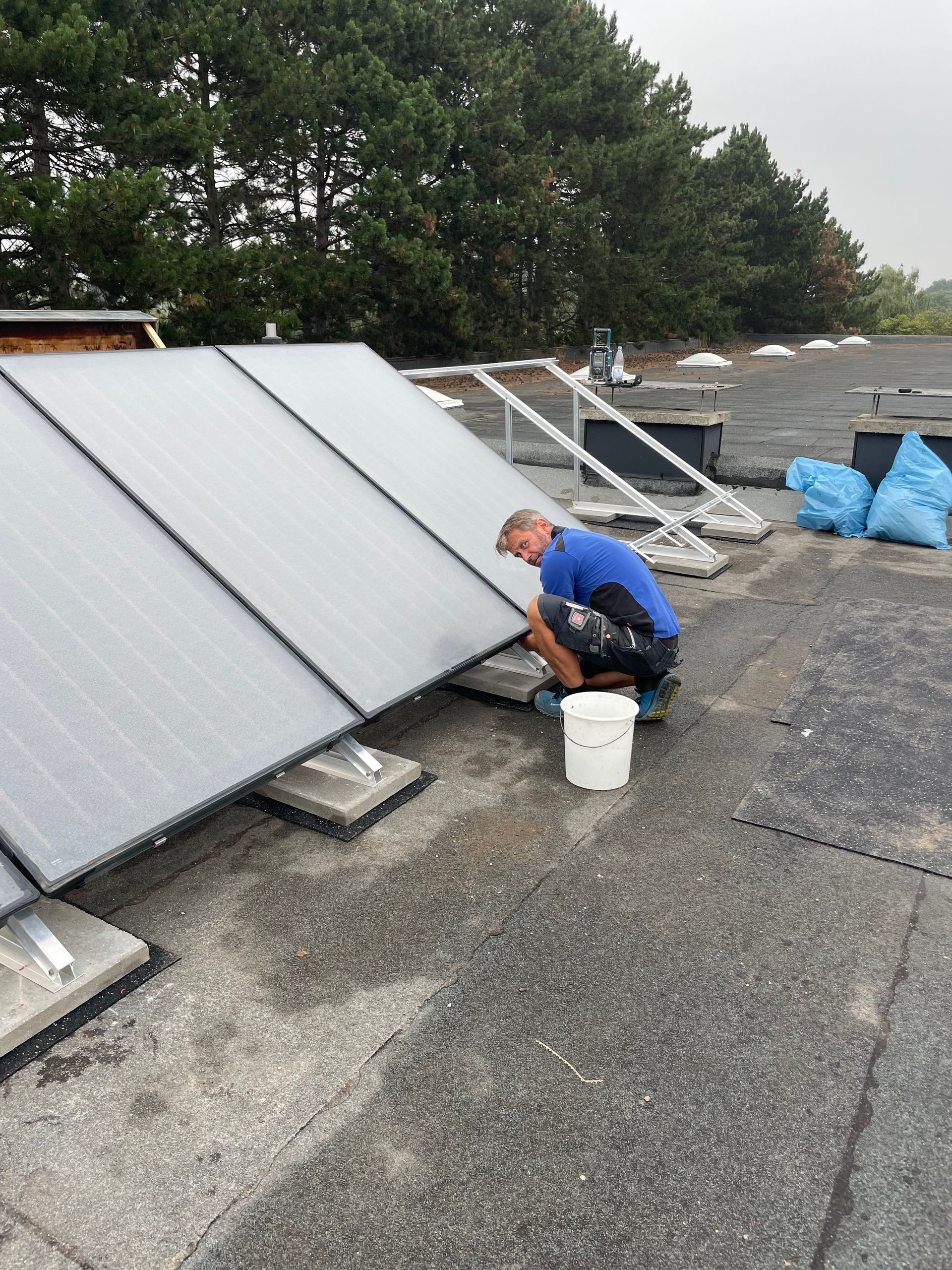 Eine Person in einem blauen Hemd arbeitet an Solarmodulen auf einem Dach. Ein Eimer mit Werkzeugen steht daneben.